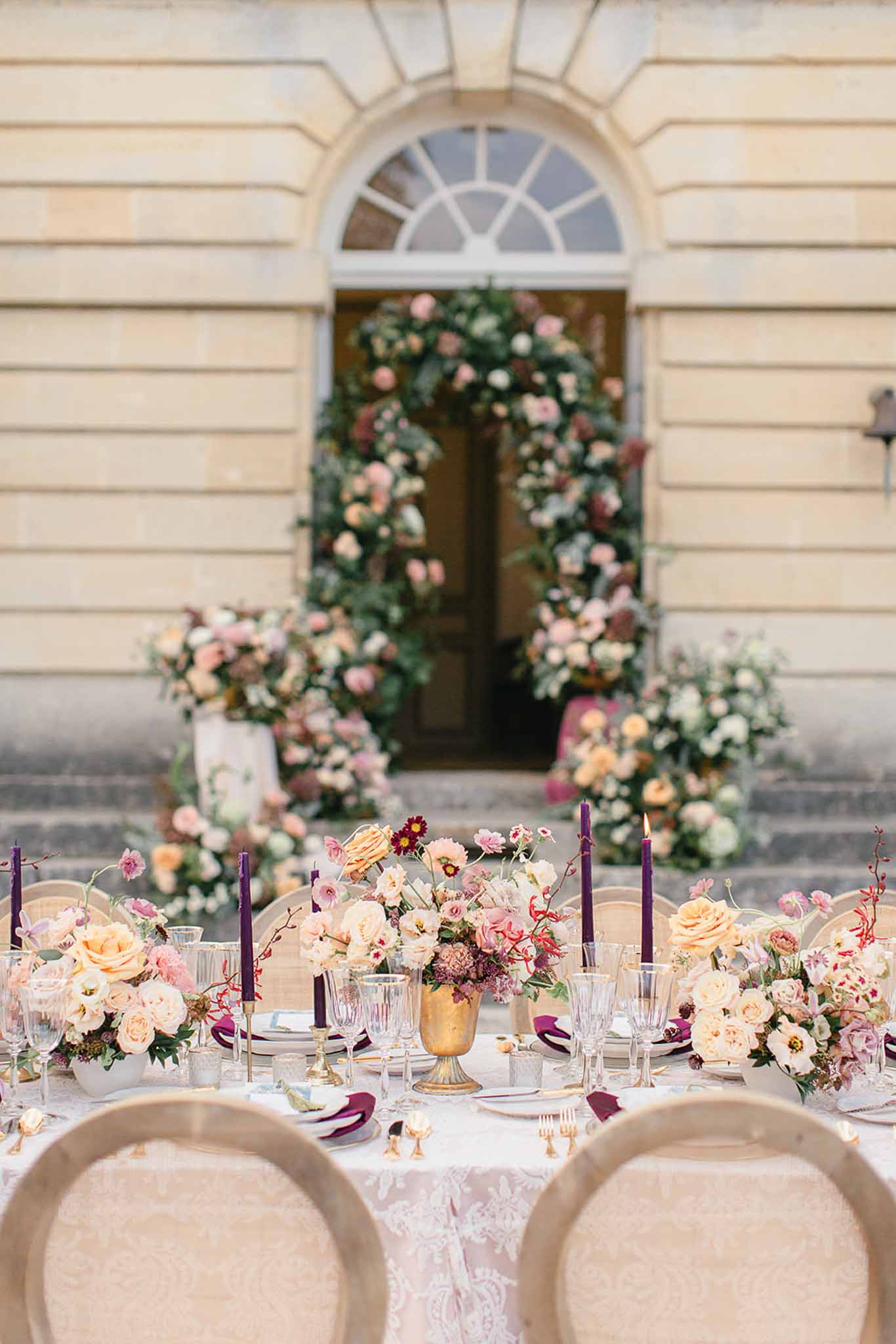 An outdoor reception tablescape set up in front of a classical French château facade, featuring a long table dressed with a white lace table runner and gold flatware. The centerpieces consist of low arrangements of peach garden roses, blush pink blooms, burgundy dahlias, and red-branched florals in gold urn-shaped vessels, interspersed with tall deep purple taper candles in brass candlesticks. Place settings include crystal glassware, gold-rimmed plates, and deep plum folded napkins. In the background, a large floral arch frames the château's arched doorway, constructed from blush pink roses, peach blooms, ivory florals, and greenery, with additional ground-level floral clusters cascading down the entrance steps. The overall color palette combines peach, blush, plum, and gold with a classic French architectural backdrop. Wide shot taken from a low angle behind two beige upholstered chairs. Potential venue feature image.