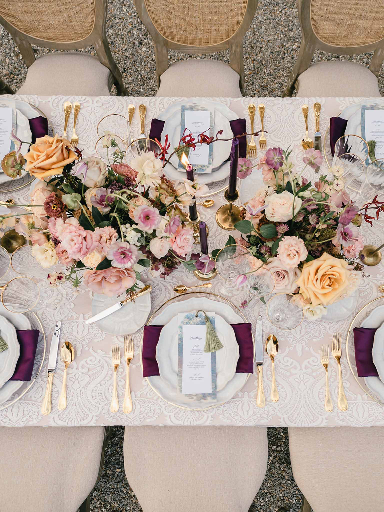An overhead flat-lay shot of an outdoor wedding reception tablescape set on a gravel surface. The rectangular table is covered with a blush and ivory damask-patterned linen and seats approximately eight guests, with cane-back chairs in a natural wood finish. Place settings feature white scalloped-edge plates layered on gold-rimmed chargers, gold flatware, clear crystal wine glasses with gold rims, deep plum/aubergine cloth napkins, and individual printed menu cards with olive green tassel ties. Two lush floral centerpieces run along the center of the table, composed of blush garden roses, mauve ranunculus, peach roses, dusty pink peonies, lavender lisianthus, burgundy orchid stems, and mixed greenery. Between the arrangements, two dark purple taper candles sit in brass candlestick holders, one lit. The overall decor palette combines blush, mauve, aubergine, peach, and gold, creating a rich, jewel-toned romantic aesthetic.