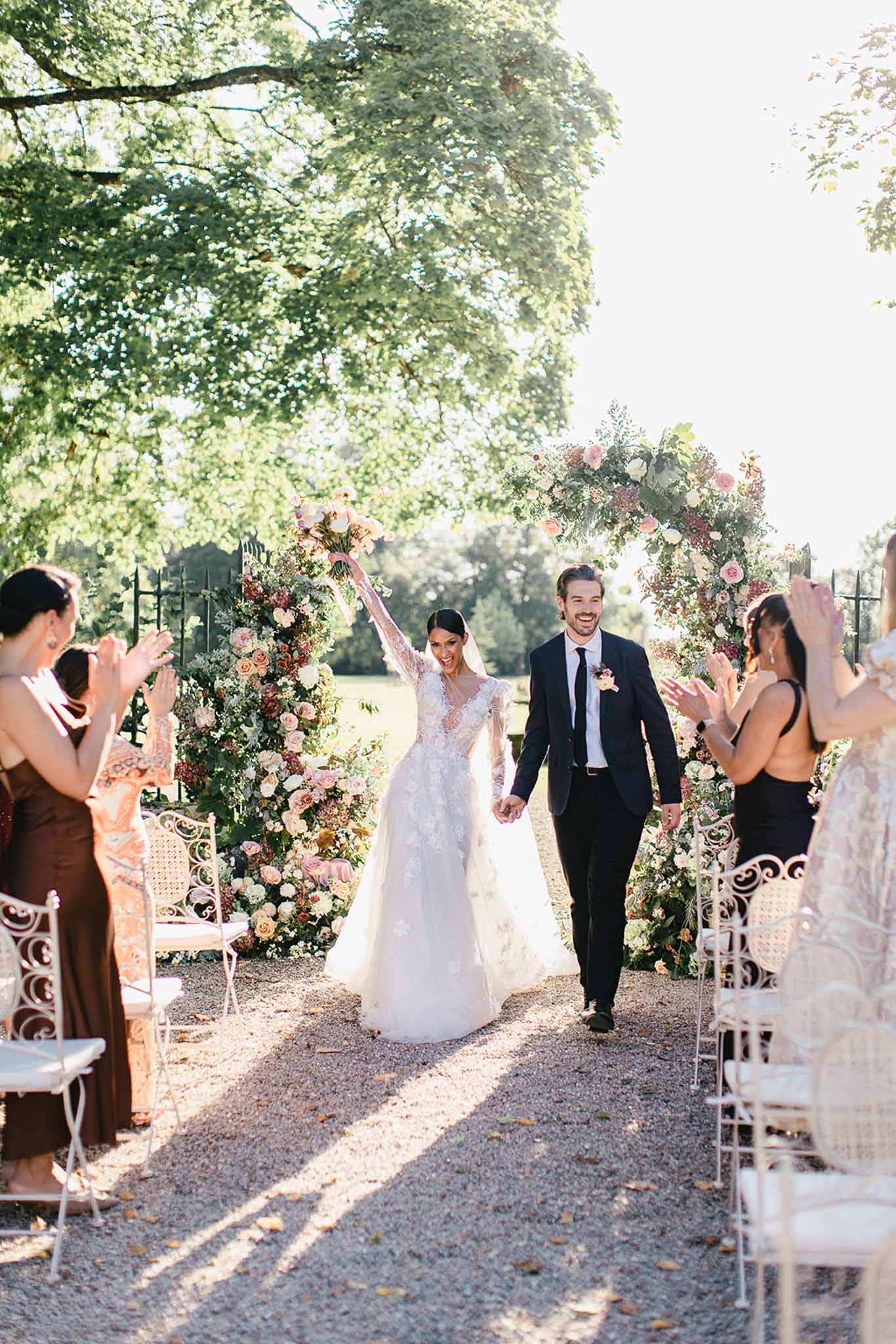 The bride and groom are walking back down the aisle together just after completing their outdoor ceremony, both smiling broadly as guests on either side applaud. The ceremony took place on a gravel path lined with white ornate wrought-iron chairs, framed by a large floral arch behind them featuring blush, dusty rose, and ivory roses, dahlias, and lush greenery. The bride wears a long-sleeve lace appliqué ballgown with a sheer bodice and holds a blush and warm-toned bouquet raised above her head in celebration, while the groom wears a navy suit with a black tie and a floral boutonnière. Guests visible on both sides are dressed in earth tones including a rust-colored slip dress and a backless black dress, contributing to an autumnal warm-toned color palette. The image is a mid-distance portrait-style shot taken in bright natural daylight with strong directional sunlight casting long shadows across the gravel path.