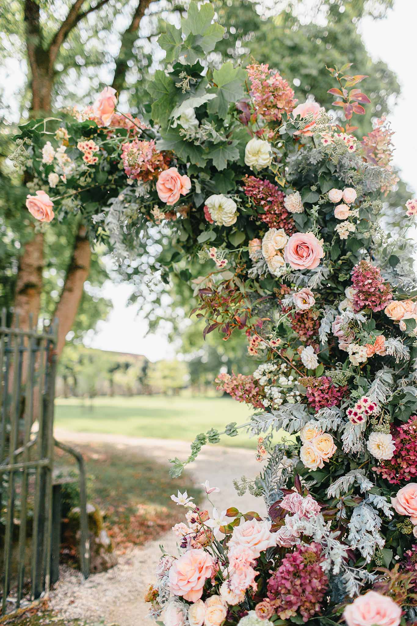 Close-up detail shot of a large outdoor floral installation, likely a ceremony arch or entrance feature, positioned along a gravel pathway with open grounds visible in the background. The arrangement is densely layered with peach and blush garden roses, cream dahlias, dusty mauve hydrangeas, small burgundy accent blooms, silver-leafed dusty miller, and abundant mixed greenery including large-leafed foliage and trailing vines. The color palette combines warm peach and blush tones with cooler mauve and burgundy shades against silvery-green foliage, creating a garden-style, romantic floral design. A dark metal gate or railing is partially visible to the left side of the frame.