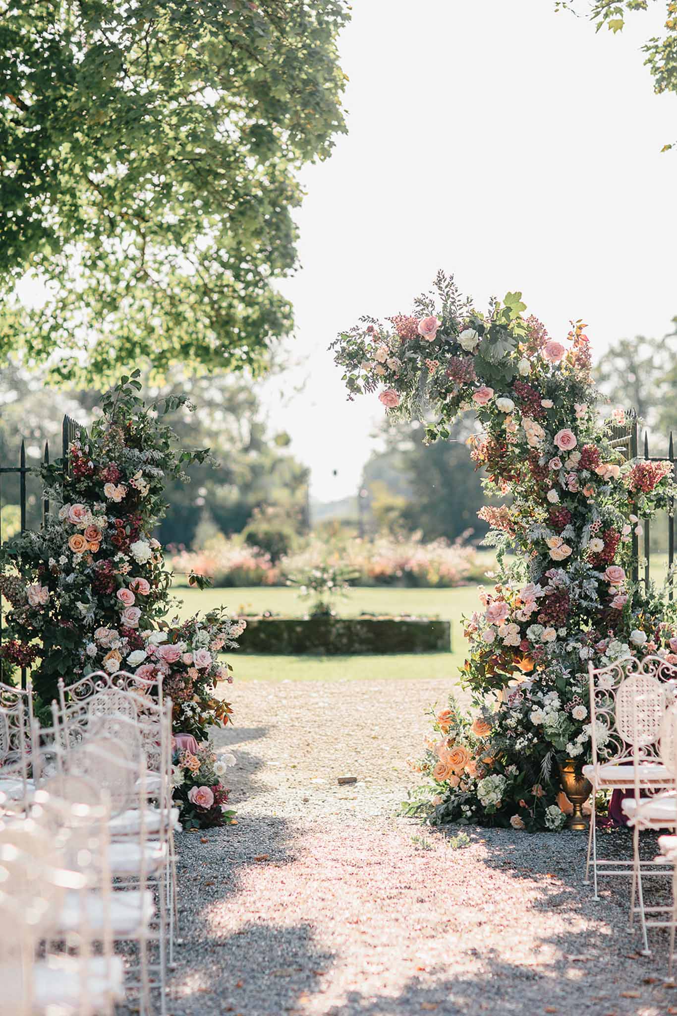 An outdoor wedding ceremony setup shot from the aisle looking toward a large circular floral arch positioned at the far end of a gravel path. The arch is densely covered in blush pink roses, peach roses, ivory garden roses, burgundy blooms, and lush mixed greenery including eucalyptus and trailing foliage. Additional floral installations extend down the base of the arch on both sides, continuing the same color palette of blush, peach, ivory, and deep burgundy. A gold urn accent is visible at the base of the right-hand installation. White ornate metal chairs with pale pink cushions are arranged in rows on either side of the gravel aisle, with rose petals scattered across the path. The setting is a formal outdoor garden with manicured hedging and a long garden vista visible through the arch. The overall styling is romantic and garden-inspired with a warm, soft color palette. Wide shot taken from ground level looking down the ceremony aisle.