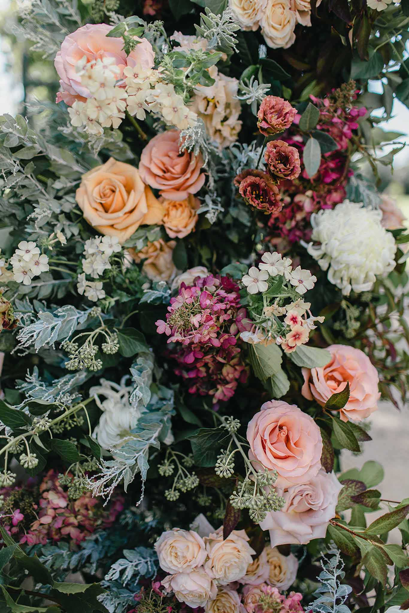 A close-up detail shot of a large floral installation, likely a ceremony arch or statement arrangement photographed outdoors. The arrangement features a rich mix of peach and apricot garden roses, blush pink roses, cream roses, burgundy lisianthus, mauve and deep pink hydrangeas, small cream phlox-like filler flowers, and white snowball hydrangeas, all woven through with lush dark green foliage, dusty miller with silver-grey leaves, and trailing ivy. The overall color palette spans warm peachy-coral tones to deep berry and burgundy, with cream accents providing contrast. The composition is a tight vertical crop that fills the frame entirely with blooms and greenery, with no people visible.