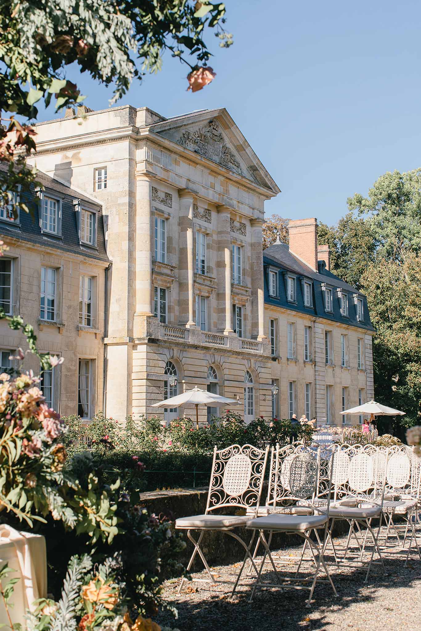 An outdoor wedding ceremony setup in the grounds of a French classical château, featuring a large neoclassical stone building with a pediment, pilasters, decorative bas-relief, and mansard roof wings. White ornate wrought-iron chairs with cream cushions are arranged in rows on a gravel terrace facing the château. Two cream parasols are open near the building's facade, and a small group of people is visible in the background near the entrance. In the foreground, a floral arrangement with peach, dusty pink, and amber blooms with mixed greenery is partially visible, likely part of a ceremony aisle or altar decoration. The shot is a wide-angle perspective framed by foliage in the upper corners, taken in bright daylight. Potential venue feature image.