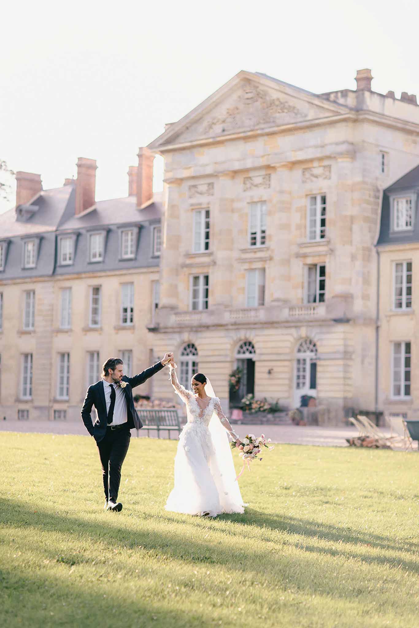 A couple portrait shot outdoors on the grounds of a French château, with the stone classical-style manor building prominently visible in the background. The groom, wearing a dark navy suit with a black tie and a small buttonhole, raises the bride's hand as she twirls on the lawn; the bride wears a long-sleeved lace appliqué ball gown with a deep V-neckline, a full ivory tulle skirt, and a cathedral-length veil, and carries a loose bouquet of blush and coral flowers with trailing ribbon. The image is a wide shot taken in warm golden-hour light, with long shadows cast across the lawn. Potential venue feature image.