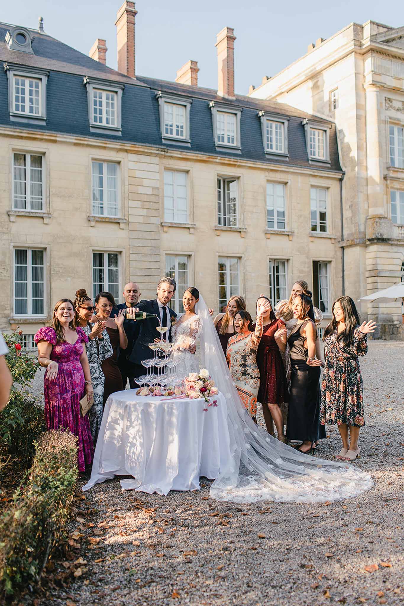 The couple is pouring champagne into a coupe tower during an outdoor cocktail hour at a French château, surrounded by approximately ten guests who are laughing and reacting with excitement. The groom, dressed in a dark suit, pours from a champagne bottle while the bride wears a long-sleeved lace wedding gown with a cathedral-length lace-trimmed veil that trails across the gravel courtyard. The champagne tower sits on a white satin-clothed round table decorated with blush and burgundy floral arrangements including what appear to be roses. Guests are dressed in a mix of colorful outfits including a fuchsia maxi dress, a dark burgundy sequined dress, a black satin midi skirt, and a patterned boho-style dress, giving the group a relaxed, festive feel. The shot is a wide portrait taken in warm late-afternoon golden light, with the classical limestone château facade featuring mansard roofing and symmetrical windows serving as the backdrop. Potential venue feature image.