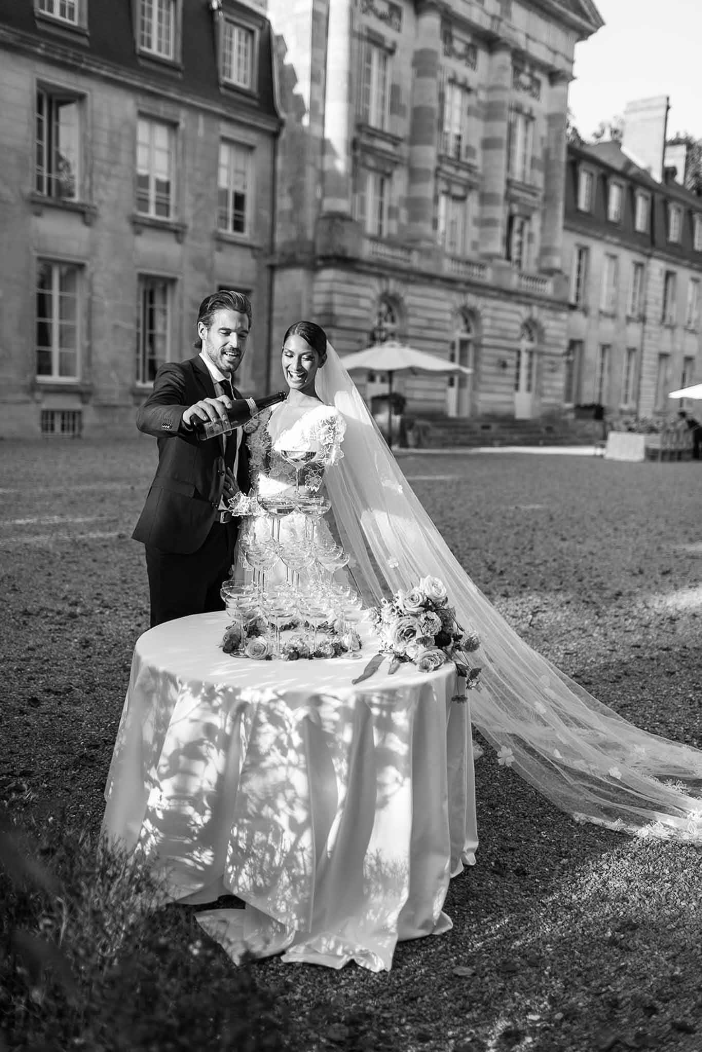 This black-and-white image shows a couple performing a champagne tower pour during what appears to be an outdoor cocktail hour or reception moment. The groom, dressed in a dark suit with a tie, pours champagne from a bottle into the top of a multi-tiered tower of coupe glasses arranged on a round table with a white floor-length linen. The bride wears a strapless lace-detailed gown and a long cathedral-length veil with floral appliqué detail that trails behind her; she is smiling broadly. The champagne tower base is decorated with what appear to be full garden roses and foliage arrangements. The setting is an outdoor courtyard or grounds of a grand French classical-style building with multi-pane windows and ornate stone façade visible in the background, along with white parasols suggesting a cocktail reception setup. The image is shot in medium portrait format with both figures fully visible and the architecture providing context in the background.