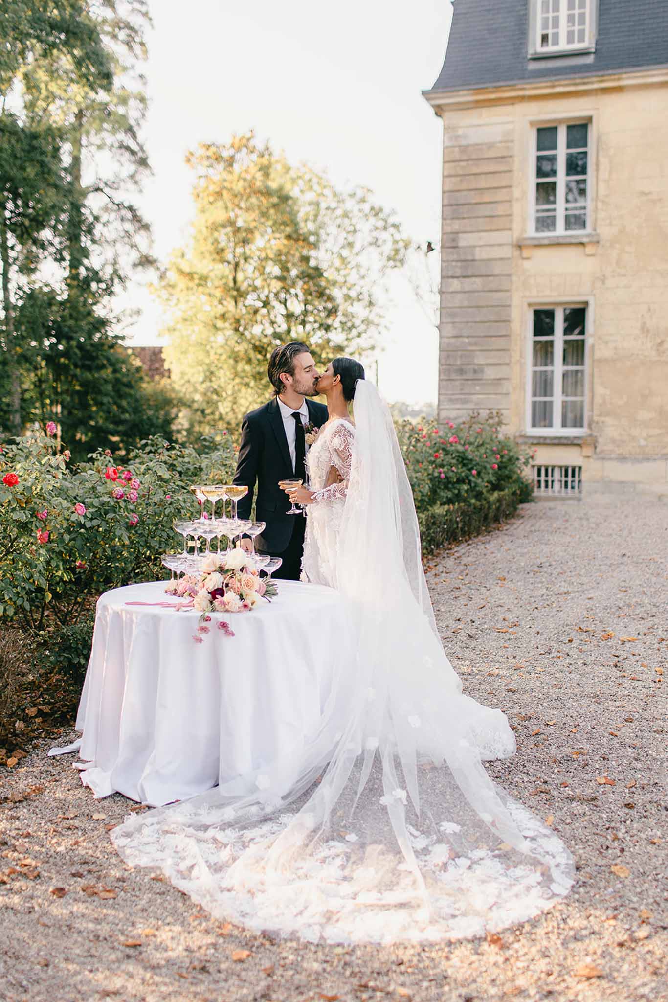 A bride and groom share a kiss outdoors on a gravel courtyard beside a champagne tower — a pyramid of coupe glasses arranged on a round table draped in a white cloth, accompanied by a bouquet of blush pink, cream, and burgundy roses and peonies. The groom wears a dark navy suit with a tie, while the bride wears a long-sleeve lace gown with intricate embroidery and a cathedral-length veil with floral appliqué trim that trails across the gravel. Both are holding champagne coupes. The shot is taken from a medium-wide angle from behind and to the side, emphasizing the full length of the veil and the table styling. The setting is the grounds of a French château with a pale stone façade visible to the right and rose bushes in bloom in the background, suggesting a classic French estate wedding aesthetic. Potential venue feature image.