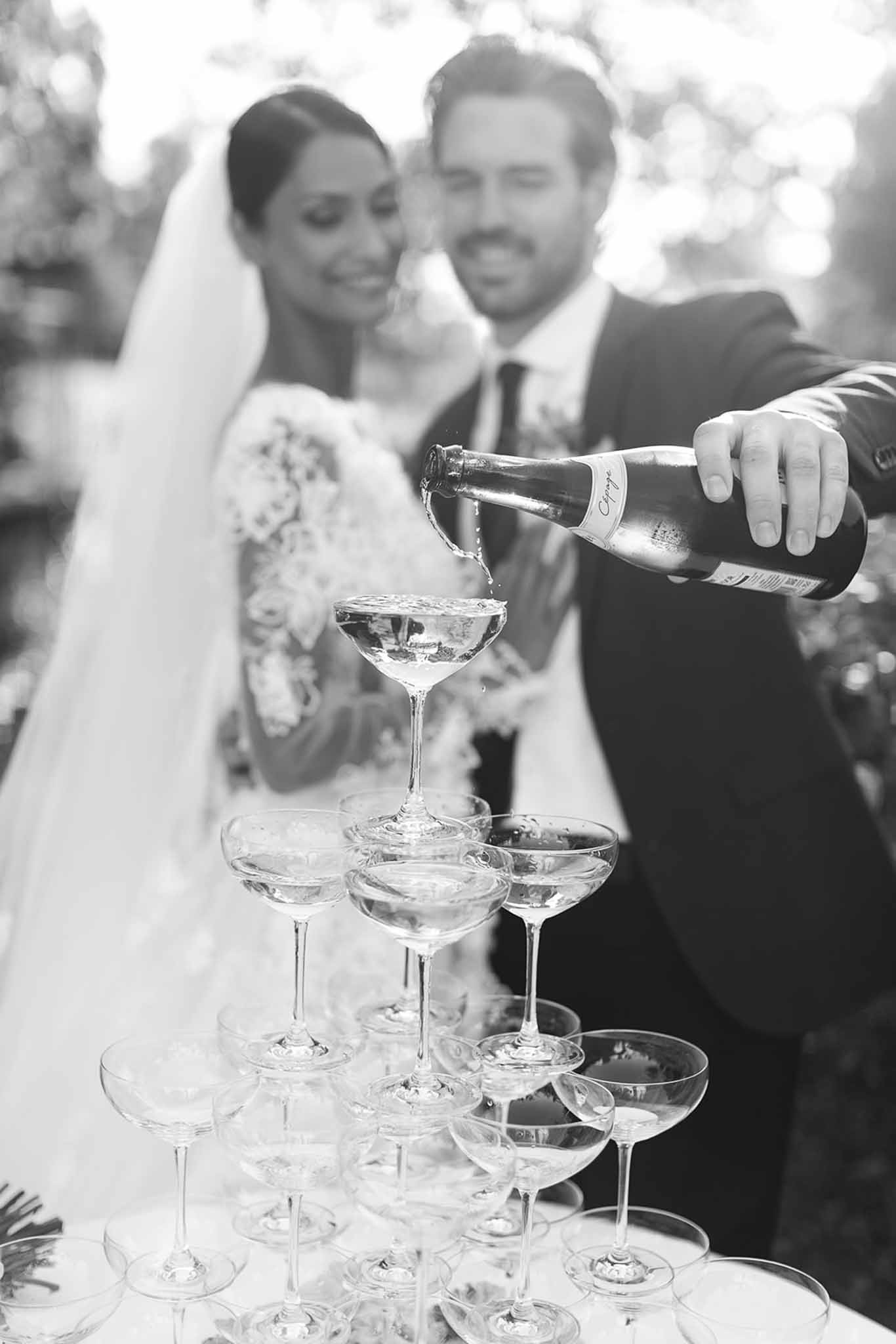 The couple is pouring champagne into a coupe glass tower during what appears to be a reception or cocktail hour, set outdoors with soft bokeh foliage in the background. The image is black and white, with strong contrast between the bright whites of the bride's long-sleeved lace gown and cathedral veil and the dark tones of the groom's suit and tie. The champagne tower consists of approximately four tiered rows of coupe glasses, with champagne visibly overflowing from the top glass down. The composition uses a shallow depth of field, keeping the tower and the pouring bottle sharp in the foreground while the smiling couple is softly blurred in the background, creating a layered portrait-style shot.