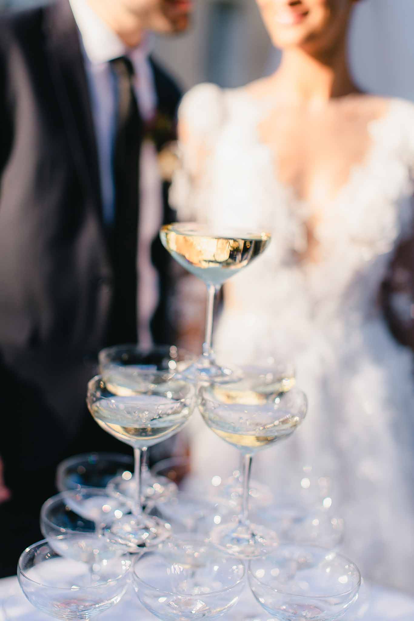 A close-up detail shot of a champagne tower made from coupe glasses filled with sparkling wine, photographed during what appears to be a cocktail hour or reception celebration. The upper tiers of the tower are in sharp focus, showing the golden champagne, while the lower glasses are softly blurred in the foreground. In the soft-focus background, the couple is visible — the groom in a dark suit with a grey tie and a boutonnière, and the bride wearing a white dress with puffed sleeves and a deep V-neckline with textured fabric detailing. The shot is taken outdoors in warm natural light, with a portrait-style composition emphasizing the tower as the primary subject.