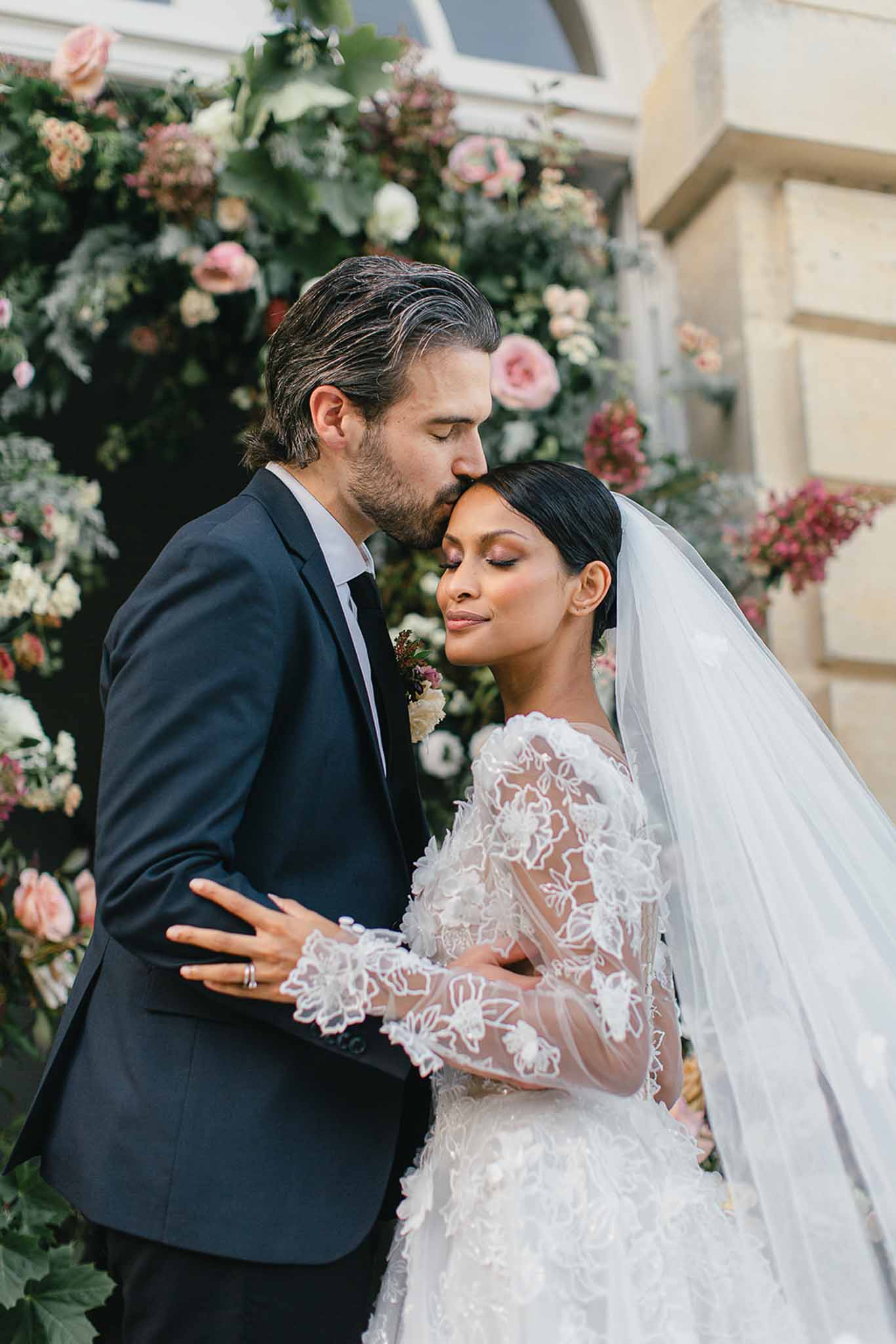 A couple portrait taken outdoors in front of a stone building facade, with the groom kissing the bride on the temple. The groom wears a navy suit with a white shirt and a dark tie, with a small floral boutonniere featuring ivory and burgundy blooms. The bride wears an ivory gown with sheer, long lace sleeves featuring floral appliqué detailing and a full tulle skirt, paired with a long cathedral-length veil worn over a sleek low bun. A large floral installation fills the background, composed of blush pink garden roses, ivory ranunculus, dusty rose blooms, burgundy florals, and lush greenery arranged against the building's entrance. The shot is a close-up portrait framed from the waist up, with soft natural light and shallow depth of field keeping focus on the couple's faces.