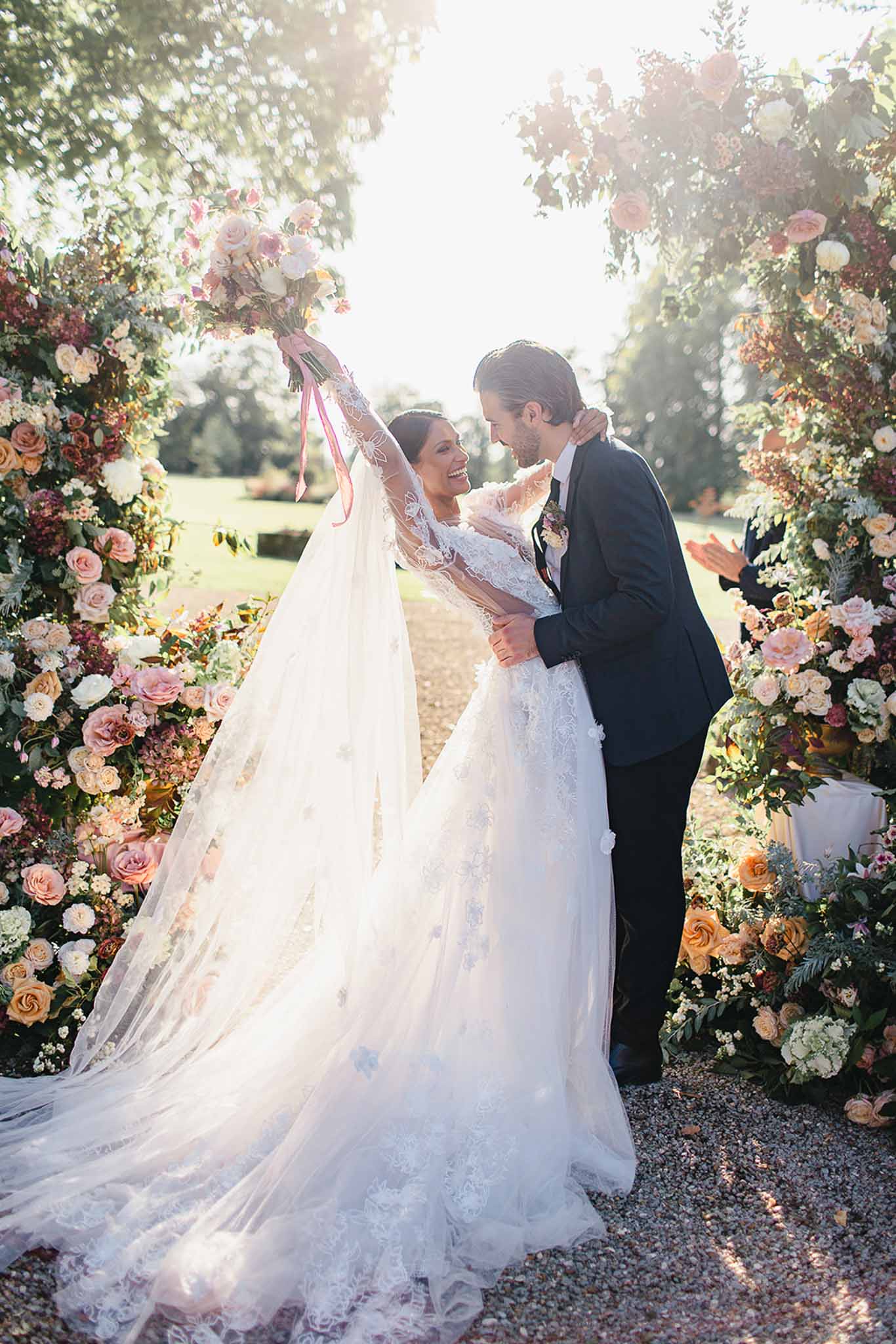 A couple celebrates just after their outdoor ceremony, standing together beneath a large floral arch in a park or garden setting with a gravel path. The bride wears a full-length white lace ball gown with long lace sleeves, floral appliqué detailing, and an extended cathedral train, while holding her bouquet of blush pink, cream, and peach roses with a trailing pink ribbon raised above her head. The groom wears a dark navy suit with a floral boutonnière and has his arms around the bride as they face each other and laugh. The ceremony arch is densely arranged with blush pink, peach, burnt orange, ivory, and burgundy blooms — including roses, dahlias, and hydrangeas — interspersed with lush greenery, framing the couple on both sides. The image is a full-length portrait shot backlit by warm golden sunlight, creating a bright, warm glow around the couple.