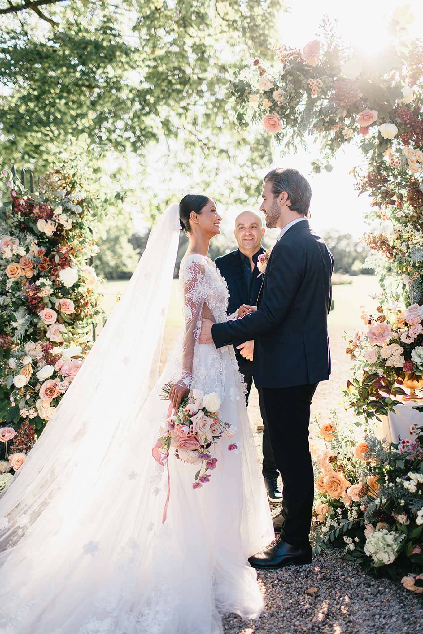 An outdoor wedding ceremony capturing the couple exchanging vows beneath a large floral arch in a garden setting with a gravel path. The bride wears a long-sleeve lace gown with sheer illusion sleeves and a full cathedral-length veil, and holds a loose bouquet of blush pink, dusty rose, and cream roses with trailing ribbons. The groom is dressed in a dark navy suit with black dress shoes. An officiant in a dark jacket stands between them. The ceremony arch is densely decorated with peach garden roses, blush and cream peonies, burgundy foliage, and trailing greenery arranged in a lush, garden-style aesthetic. Warm backlight from the sun creates a soft glow through the trees behind the arch. The shot is a medium-wide portrait framing the couple and arch from ground level.