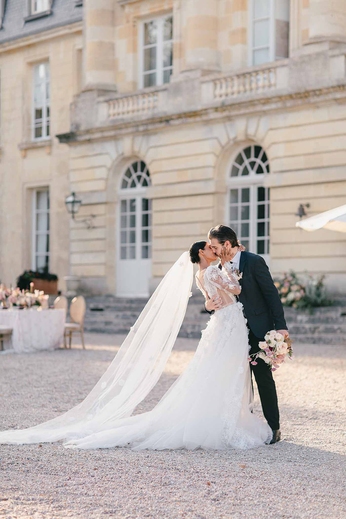A couple portrait taken outdoors on the gravel courtyard of a French château, with the bride and groom sharing a kiss. The bride wears a white long-sleeve lace gown with 3D floral appliqués, a full skirt, and a cathedral-length veil that trails across the ground; she holds a loose bouquet of blush and ivory roses with hanging garden elements. The groom is dressed in a dark navy suit. In the soft-focus background, the limestone façade of the château is visible along with a reception table set with a white linen cloth, gold chairs, and pink floral centerpieces with candles to the left. The image is a full-length portrait shot with warm late-afternoon light, classic French architecture styling, and a romantic but understated aesthetic.