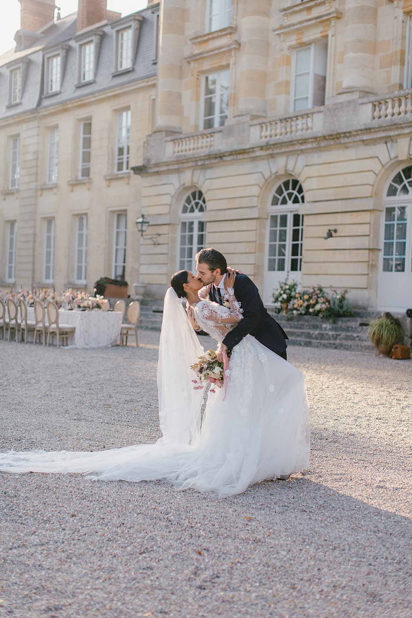 A couple shares a dip kiss on the gravel forecourt of a French château, posing for a portrait during what appears to be couples' portraits following the ceremony. The bride wears a long-sleeved lace gown with a full tulle skirt and an extended cathedral-length veil, and holds a loose bouquet featuring blush, burgundy, and warm-toned blooms. The groom is dressed in a dark navy suit. In the background, an outdoor reception setup is partially visible, with long white-linen tables, gold Louis chairs, and low floral centerpieces in peach and cream tones arranged along the château façade. The overall styling is classic French with romantic floral accents. Wide portrait shot taken in warm late-afternoon light. Potential venue feature image.