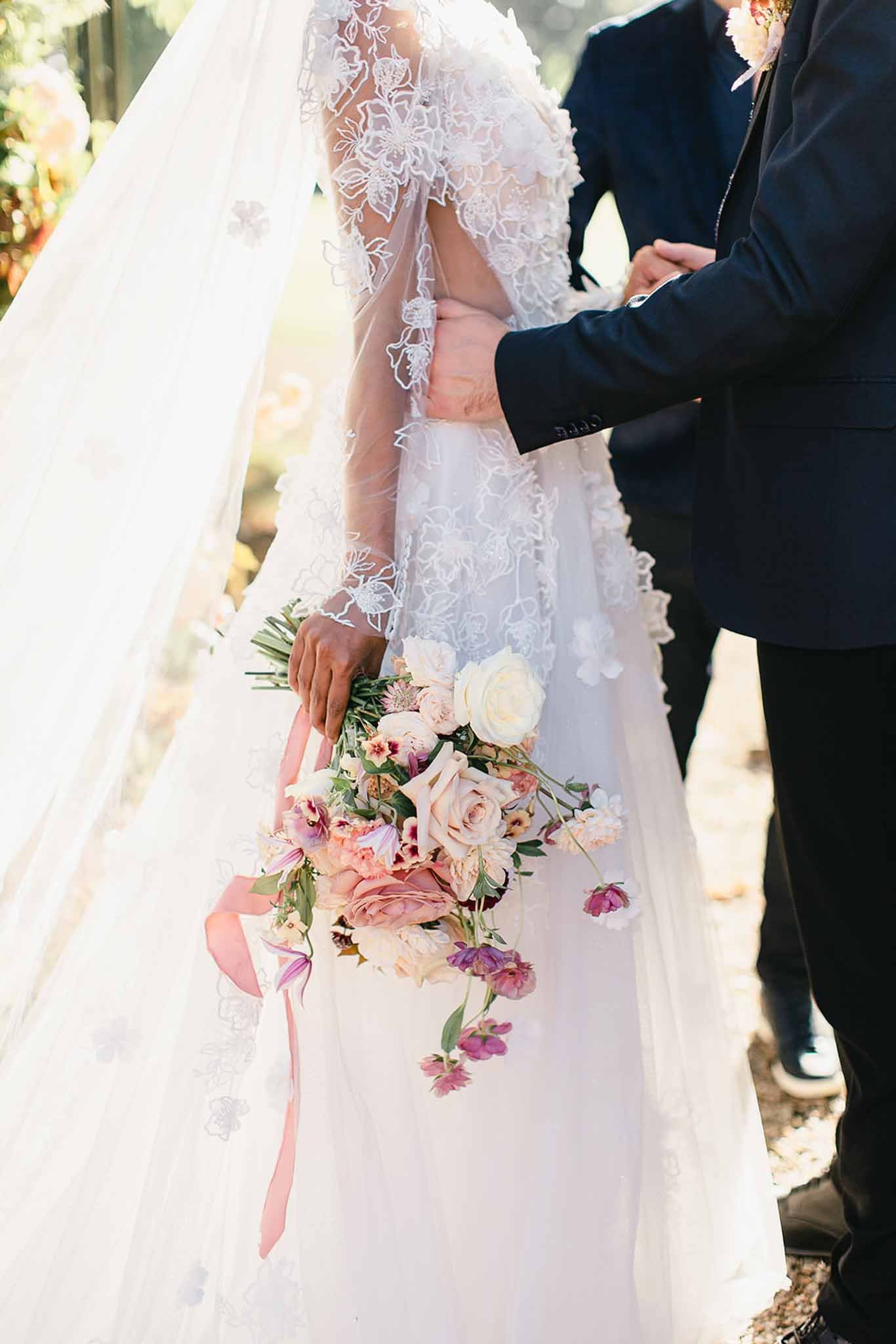 A close-up portrait of a bride and groom standing together outdoors, cropped at the torso — neither face is visible. The bride wears a white long-sleeved lace gown with intricate floral embroidery and a cathedral-length veil with matching lace appliqué edges. The groom, positioned to her right, wears a navy blue suit and has his hand placed at her waist. The bride holds a loose, garden-style bouquet at her side composed of blush roses, cream ranunculus, mauve peonies, alstroemeria in burgundy and peach tones, and small purple scattered blooms, tied with a trailing dusty pink silk ribbon. The composition is a mid-range detail shot emphasizing the dress texture, veil, and bouquet against a softly blurred outdoor background.