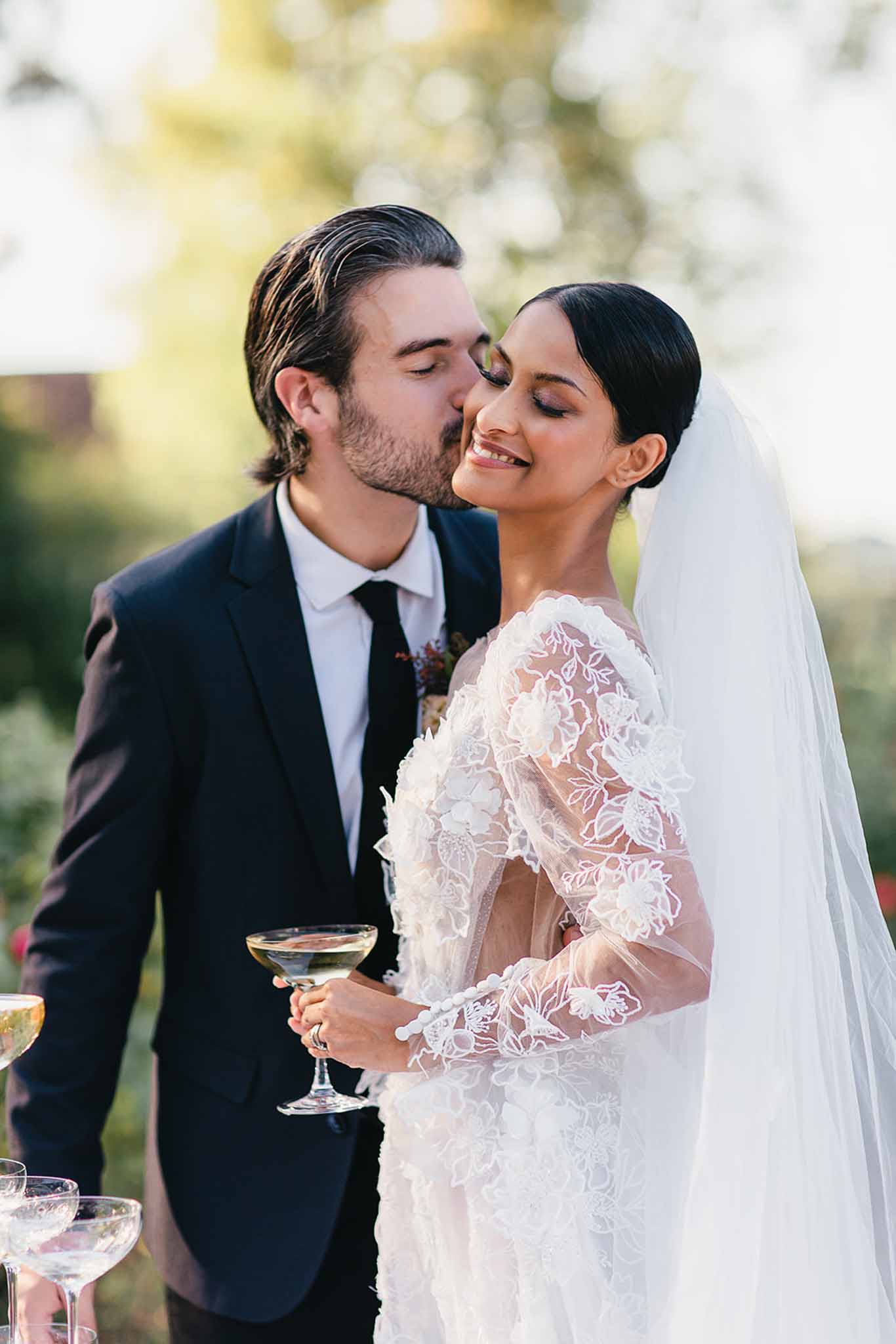 A close-up portrait of a couple during what appears to be an outdoor cocktail hour, with the groom kissing the bride on the cheek while she smiles with her eyes closed. The groom wears a dark navy suit with a black tie and a small dark floral boutonnière. The bride wears a long-sleeve white lace wedding dress with sheer floral appliqué detailing and a cathedral-length veil, with her dark hair pulled back into a sleek low bun. She holds a coupe glass filled with white wine or champagne, and additional coupe glasses are visible in the lower foreground. The background is heavily blurred with soft natural light, suggesting an outdoor garden or grounds setting. The overall styling is classic and polished.