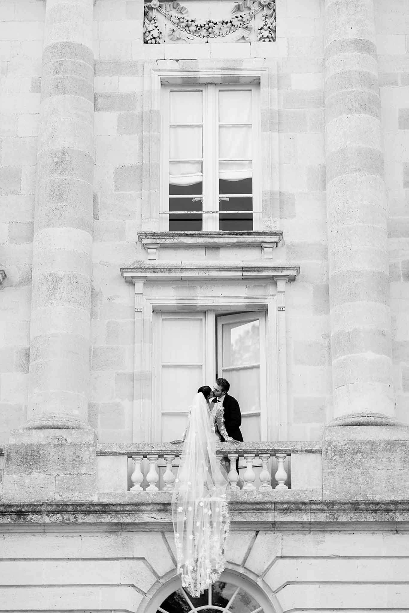 A black-and-white couple portrait taken outdoors on the stone balcony of a French château, shot from a low angle that emphasizes the grand classical architecture — fluted columns, carved stone detailing, tall French windows, and a balustraded balcony. The bride and groom are kissing at the center of the frame; the bride wears a long veil with scattered floral appliqué that drapes down over the balustrade toward the camera, and the groom is dressed in a dark suit. The composition is wide and centered, with the couple small relative to the façade, making the building's scale the dominant visual element. High contrast between the pale limestone and the darker tones of the couple's figures is characteristic of the B&W treatment. Potential venue feature image.