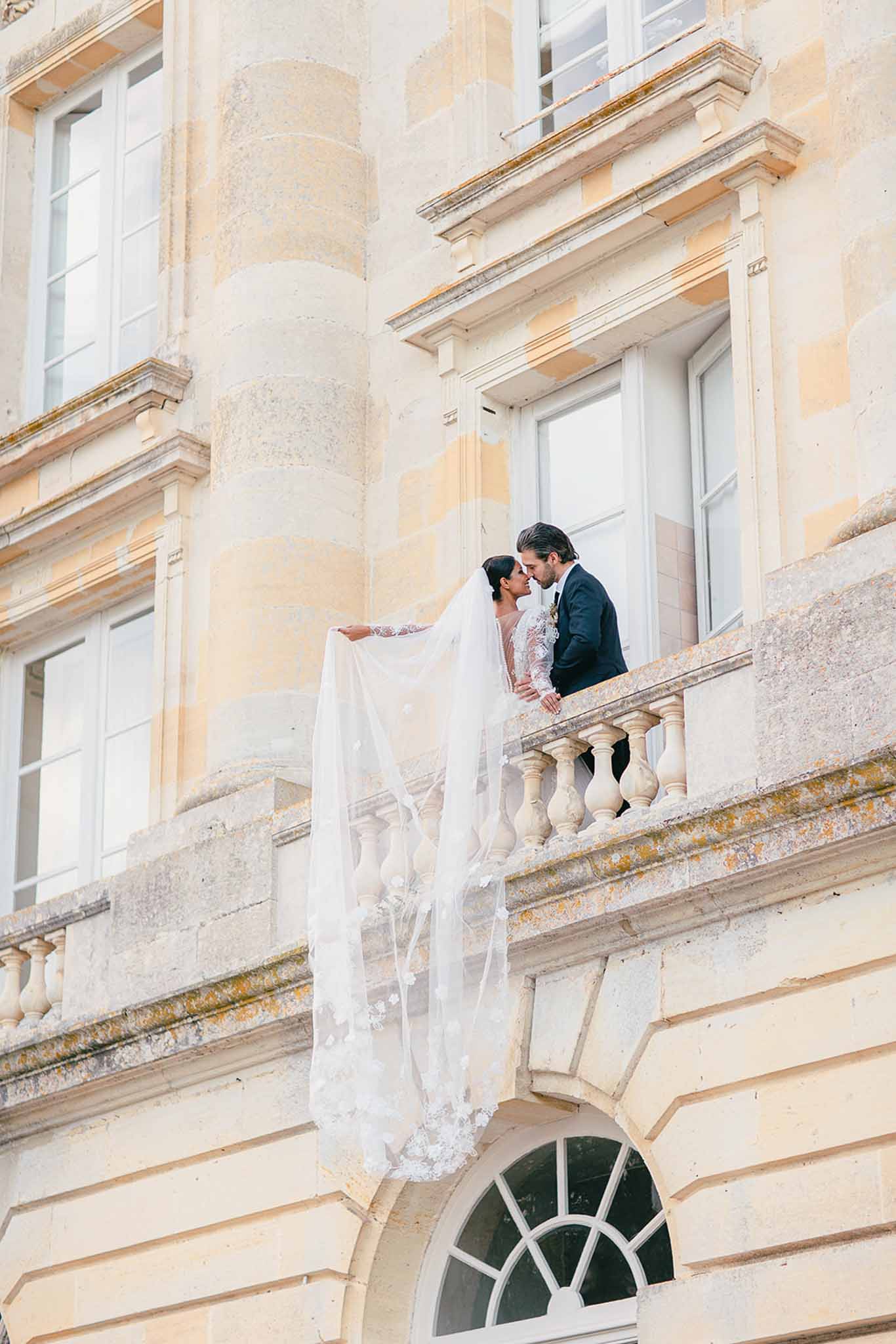 A couple portrait taken from a low angle, showing the bride and groom standing on an ornate stone balcony of a classical French château, leaning in toward each other as if about to kiss. The bride wears a long-sleeve lace gown with a cathedral-length veil embellished with 3D floral appliqués, which cascades over the balustrade and hangs down the façade of the building. The groom is dressed in a navy suit. The composition is a wide exterior shot that emphasizes the cream-toned ashlar stone façade, arched windows, and carved baluster railing of the château. Potential venue feature image.