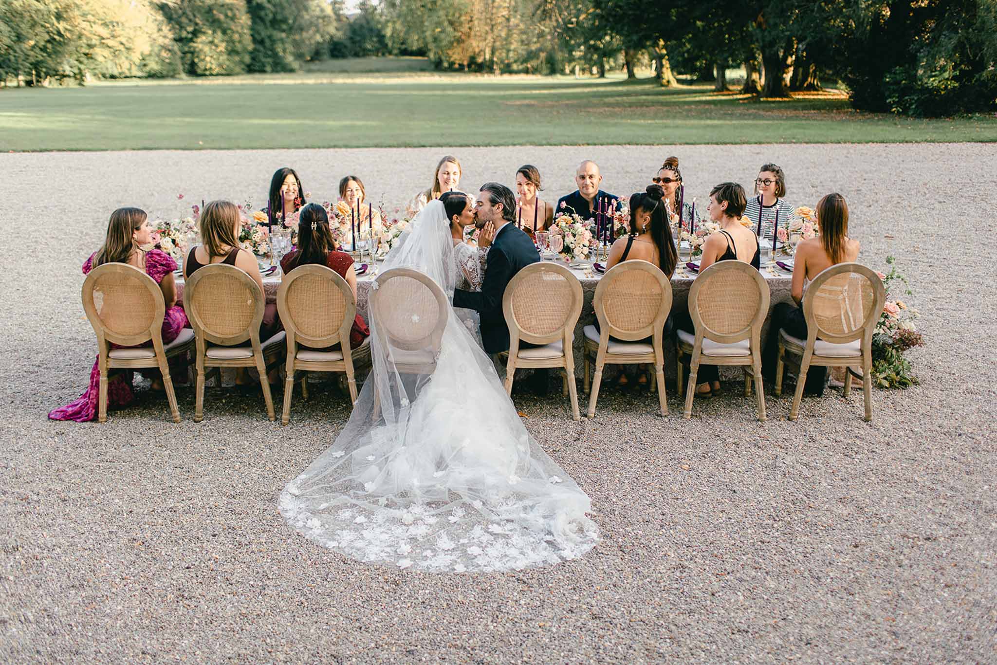 The bride and groom share a kiss while seated at the center of a long outdoor reception table on a gravel forecourt, surrounded by approximately 14 guests. The bride wears a white lace gown with a cathedral-length veil adorned with floral appliqués that trails dramatically across the gravel behind her chair; the groom wears a dark navy suit. Guests are dressed in a mix of colorful attire including deep fuchsia, burgundy, black, and patterned outfits. The rectangular dining table is set with cane-back Louis XVI-style wooden chairs and decorated with abundant loose floral arrangements in tones of blush, peach, coral, and ivory, alongside tall dark taper candles in deep purple and black candleholders, purple glassware, and what appears to be a printed or textured table covering. The wide-angle shot is taken from a slightly elevated angle looking down the length of the table, capturing the full scene including the expansive parkland setting behind the guests.
