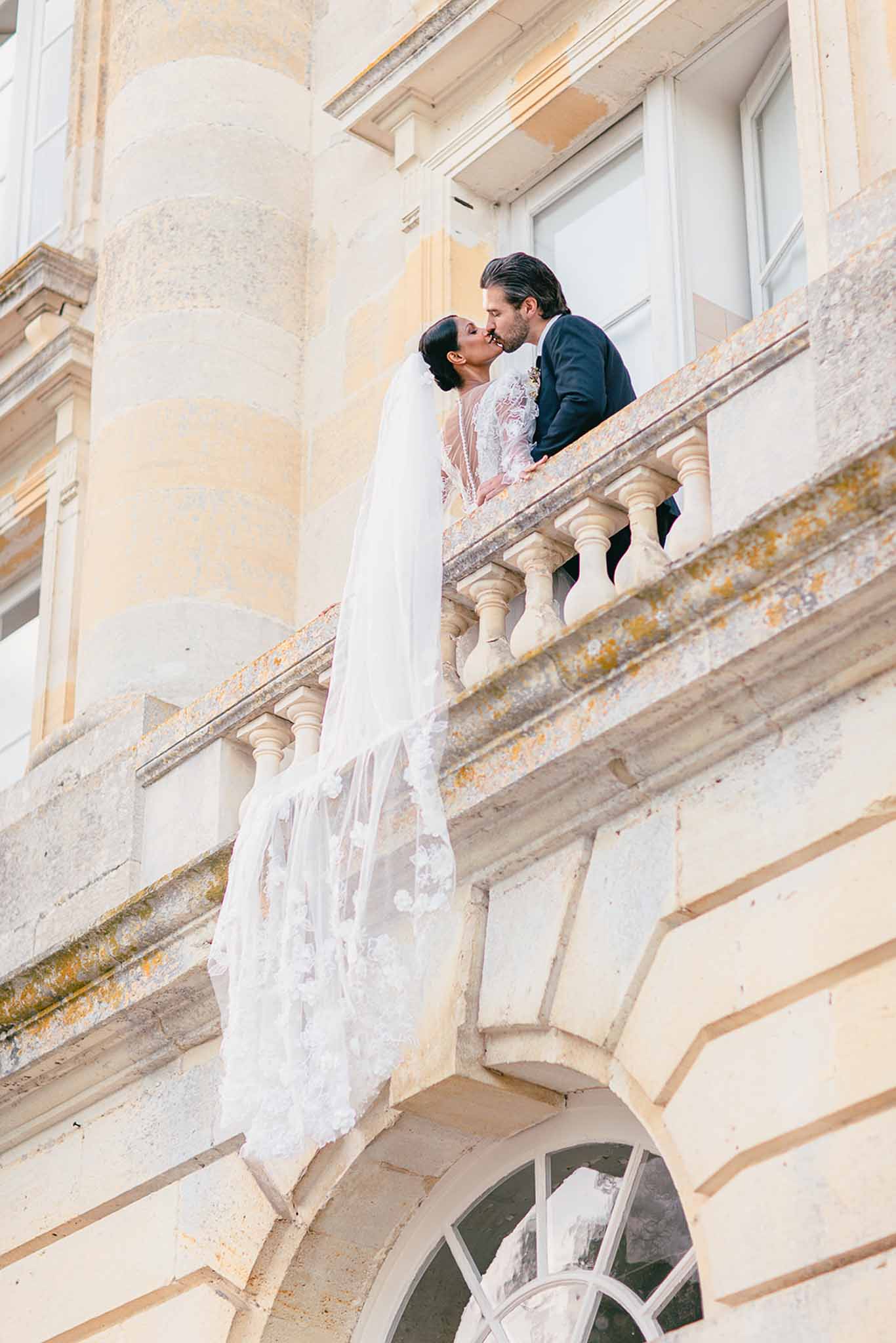 A couple shares a kiss on a stone balcony of a French château, photographed from a low angle looking up. The bride wears a long-sleeve lace gown with sheer detailing and a cathedral-length veil with floral appliqués that drapes over the balustrade and cascades down the façade. The groom is dressed in a navy suit. The architectural setting features classical balusters, arched windows, and pale stone masonry. The composition is a wide portrait shot taken from ground level, emphasizing the height of the balcony and the full length of the veil against the building's façade. Potential venue feature image.