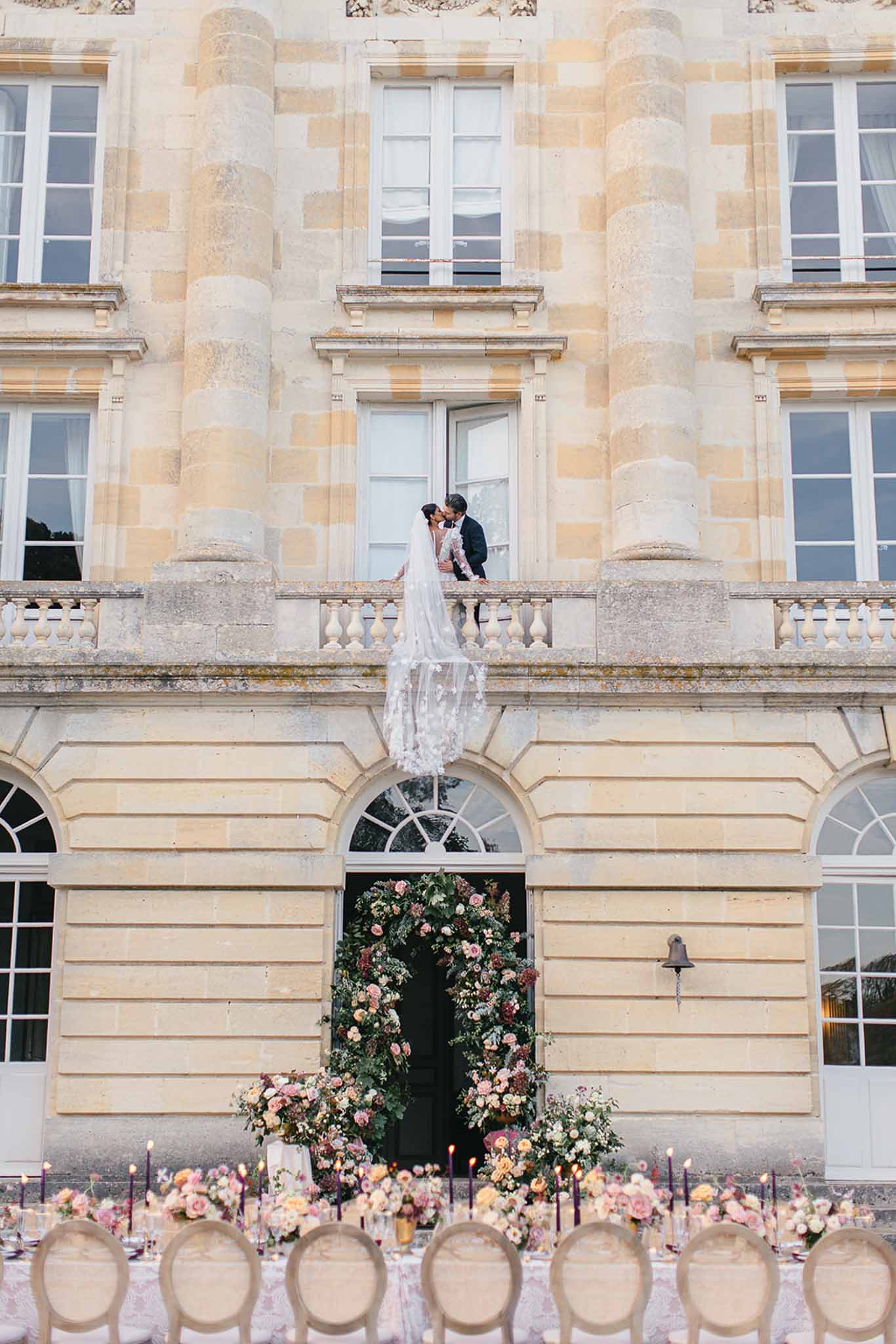 A wide outdoor shot in front of a French château, showing a bride and groom kissing on a first-floor stone balustrade balcony while the bride's long floral-appliqué veil cascades down the façade below them. The bride wears a long-sleeved white gown and the groom is in a dark suit. Below on the ground level, the château's arched entrance door is framed by a large circular floral installation composed of blush, peach, dusty rose, and deep burgundy blooms with trailing greenery. In front of the entrance, a long outdoor reception table is set with upholstered round-back chairs in natural linen tones, abundant low floral arrangements in the same blush, peach, and mauve palette, and tall deep purple taper candles running the length of the table. The classical French château architecture with ashlar stonework serves as the backdrop to the entire composition. Potential venue feature image.