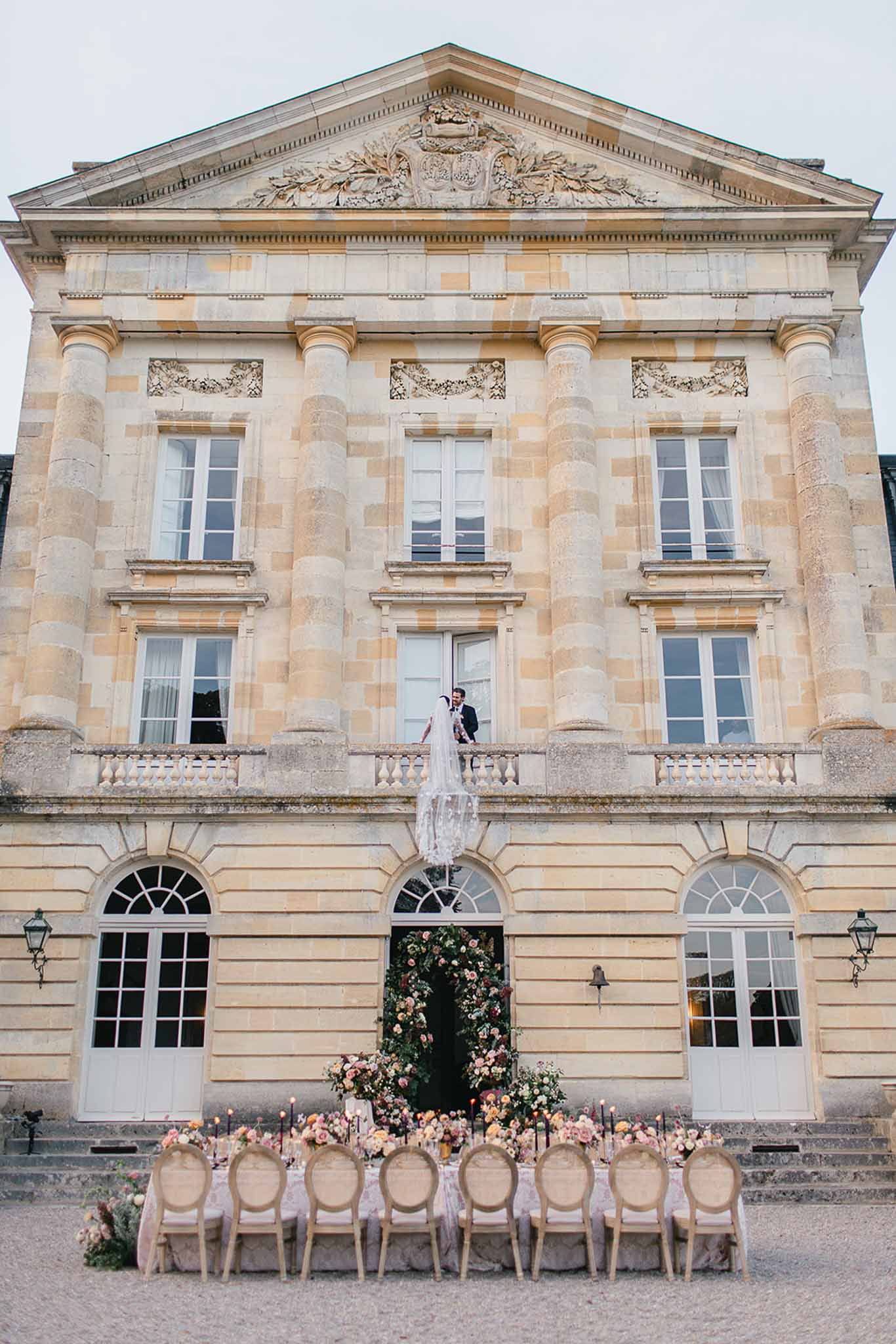 A wide shot taken from outside a French classical château, featuring a couple — a bride in a long lace gown with an extended veil cascading over the first-floor balcony railing, and a groom in a dark suit — standing on the central balcony of the building. On the ground level directly below them, a long rectangular reception table is set in front of the château's central entrance, which is framed by a large floral arch composed of blush, peach, coral, and ivory blooms with lush greenery. The table is dressed with a blush or pale pink linen and lined with French Louis XVI-style upholstered chairs in natural/greige tones. Table decor includes clusters of peach, blush, and coral floral arrangements, tall black taper candles, and what appear to be gold accent details. The overall styling palette is soft romantic with warm tones against the pale stone façade of the château. Potential venue feature image.