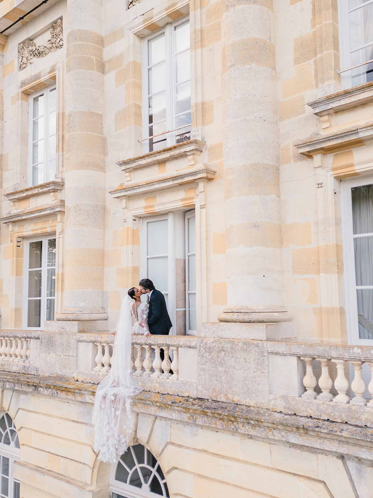 A couple shares a kiss on an ornate stone balustrade balcony of a classic French château, captured in a wide portrait shot. The bride wears a long-sleeve lace gown with a cathedral-length veil that drapes over the balcony ledge, and the groom is dressed in a dark navy suit. The façade behind them features tall French windows with white frames, carved stone detailing, and pilasters in warm cream and tan tones. The composition places the couple small within the frame, emphasizing the scale and classical architecture of the building. Potential venue feature image.