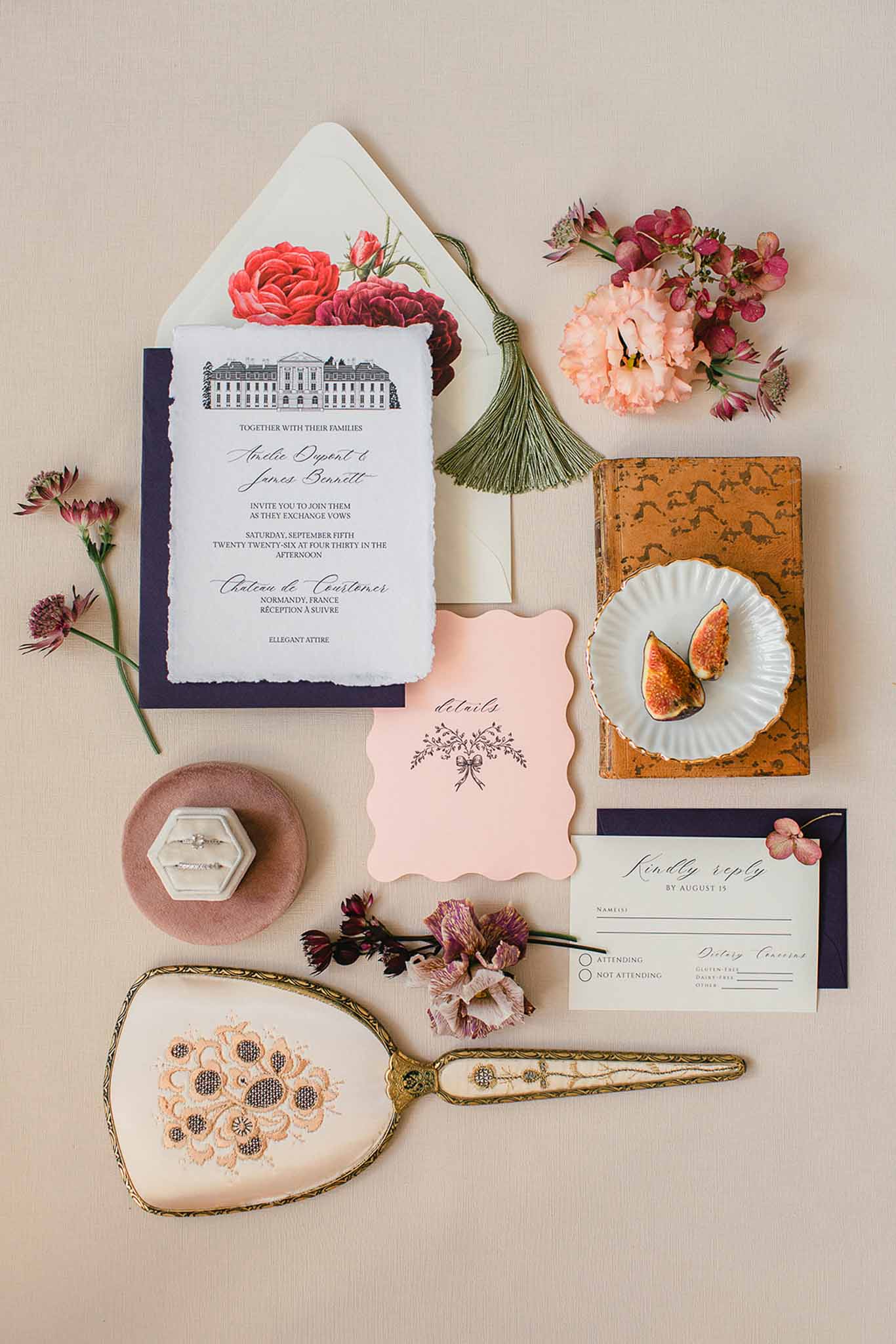 A flat lay detail shot of a wedding stationery suite arranged on a warm beige linen surface. The invitation features deckled edges on white card stock with a hand-illustrated château building at the top, layered on a navy backing card, tucked into a cream envelope lined with a printed floral pattern of red and burgundy roses, finished with a dark green silk tassel. The suite also includes a blush pink wavy-edged details card with dark script and a bow motif, a cream RSVP card with navy envelope, and a small dried floral sprig. Styling props include a vintage gold-handled hand mirror with embroidered cream and peach needlepoint back, an antique weathered book topped with a white scallop-edged dish holding two halved fresh figs, a dusty mauve velvet ring dish holding a hexagonal grey velvet ring box with a diamond engagement ring, and loose blooms in blush, burgundy, and mauve tones including astrantia and lisianthus. The overall palette is navy, blush, and warm gold with a classic French romantic styling theme.
