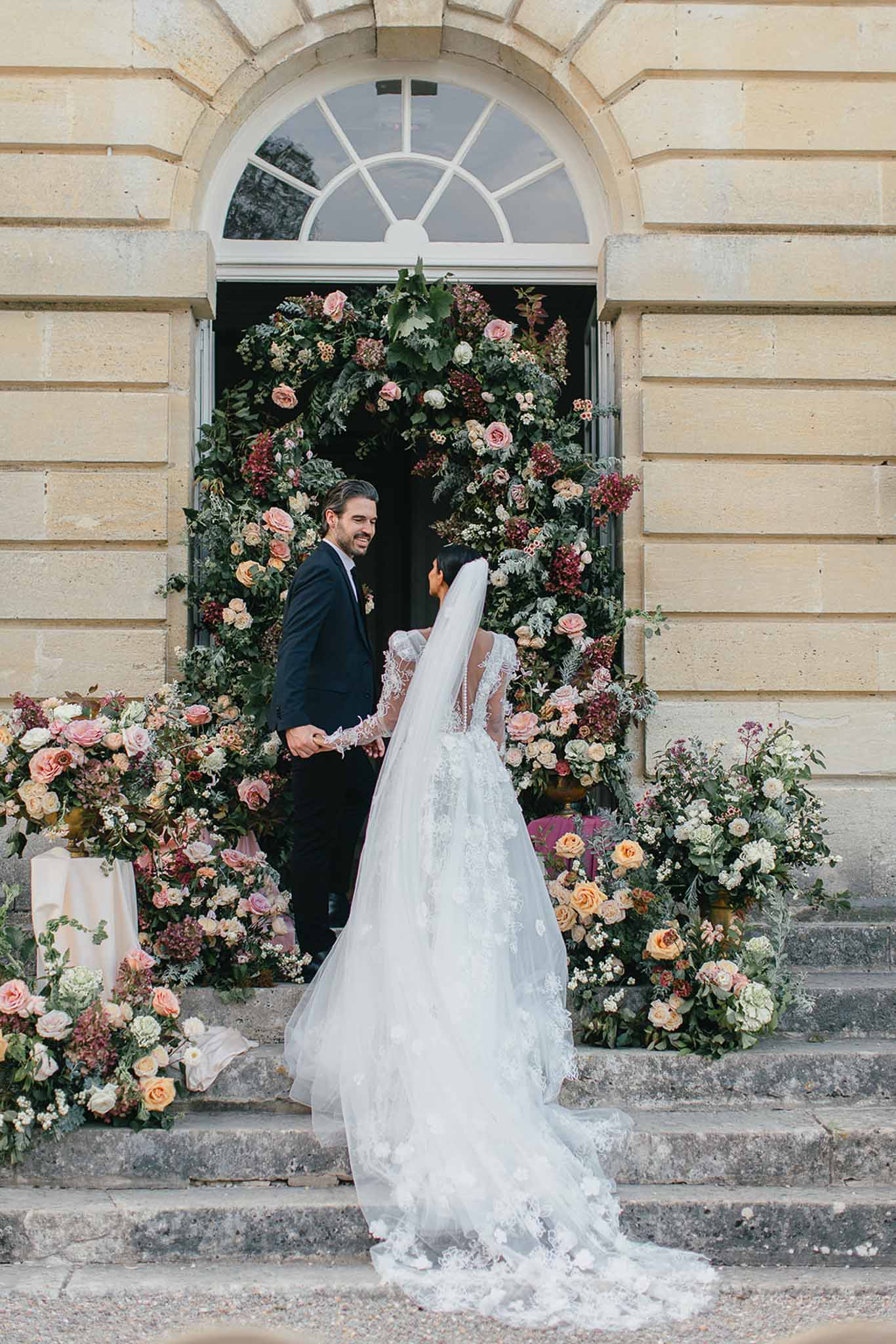A couple poses together on the stone steps of a classical French château entrance, holding hands and facing each other. The bride wears a white ballgown with long lace-embroidered sleeves, a deep illusion back, and a long cathedral-length veil with floral appliqué trim; the groom wears a navy suit with a dark tie. Framing the arched doorway behind them is a large circular floral installation composed of lush greenery, blush and apricot garden roses, deep burgundy foliage, ivory blooms, and dark red berries, with additional ground-level floral arrangements in the same rich autumn palette flanking the steps on both sides. The composition is a medium portrait shot taken from behind the bride, capturing the full length of her veil and train against the warm limestone façade. Potential venue feature image.