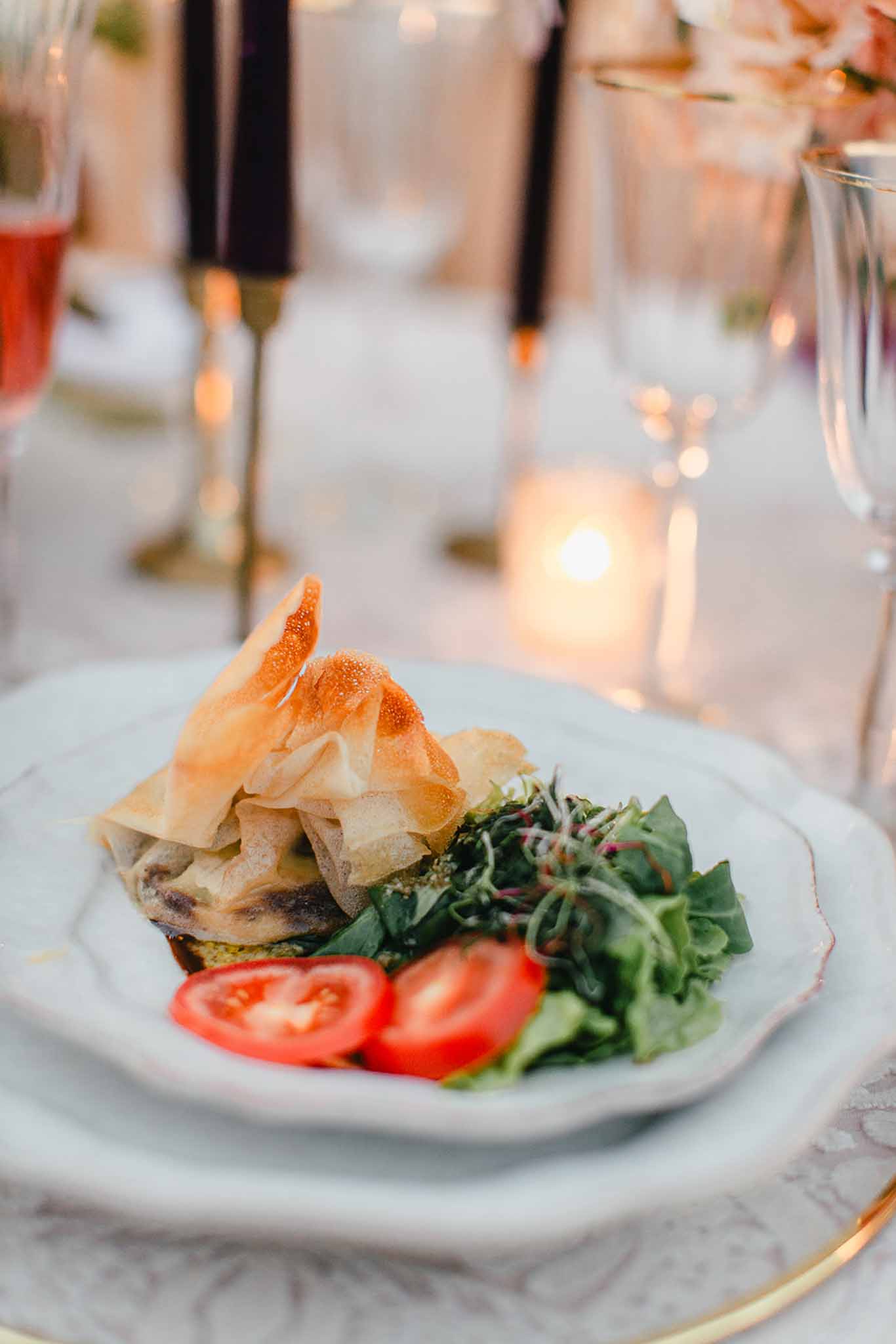 Close-up detail shot of a plated starter course at a wedding reception dinner. The white scallop-edged porcelain plate sits on a gold-rimmed charger and features a phyllo pastry parcel, a mixed green salad with microgreens, and two halved cherry tomatoes. In the soft-focus background, the table is styled with dark burgundy/black taper candles in gold candlestick holders, lit votive candles in glass holders, crystal glassware, and what appears to be a pink/blush drink, suggesting a classic yet moody reception decor palette with warm candlelight.