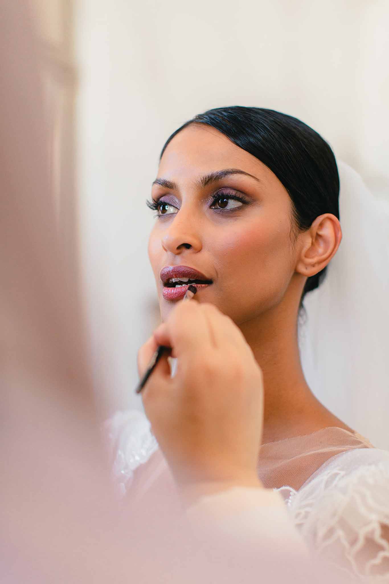 A getting-ready scene captured in a close-up portrait showing a bride having her lip liner or lipstick applied by a makeup artist's hand holding a small brush. The bride has dark hair styled in a sleek low bun, wears a white veil, and is dressed in a white gown with feather or lace detailing at the shoulder. Her makeup features a deep mauve-berry lip, warm smoky eye with a lavender-purple tone, defined brows, and voluminous lashes. The shot is taken through or around a mirror, creating a slightly soft foreground edge on the left side. The background is a plain cream-toned wall, keeping the focus entirely on the bride's face and the makeup application detail.