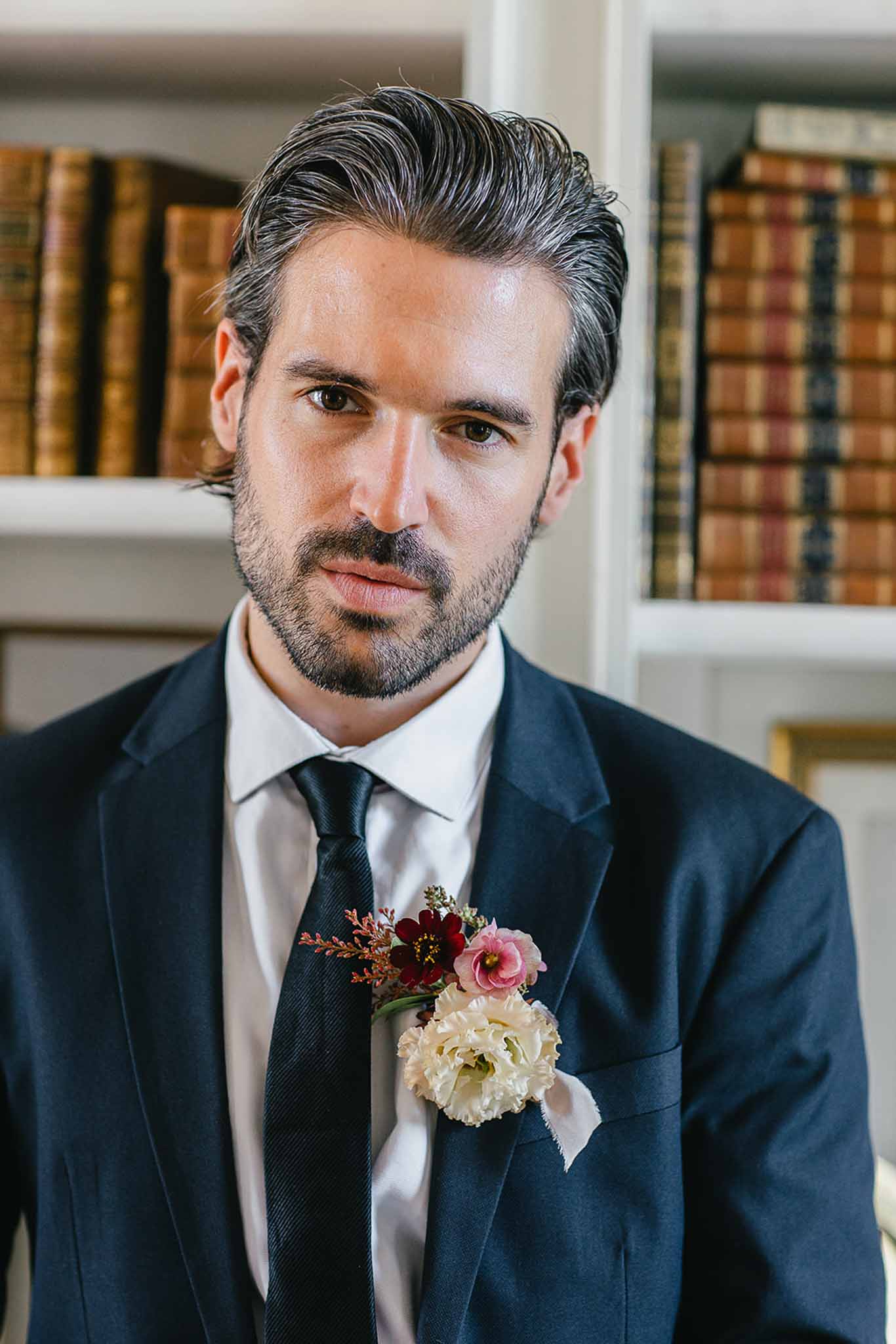 Close-up portrait of a groom wearing a navy blue suit, white dress shirt, and a dark navy tie. His boutonniere features an ivory carnation, a small pink flower, a deep burgundy bloom, and copper-toned foliage accents. He has dark, slicked-back hair and a short beard. The background shows white-painted bookshelves lined with leather-bound antique books, suggesting an indoor library or study setting. The overall styling is classic and polished.