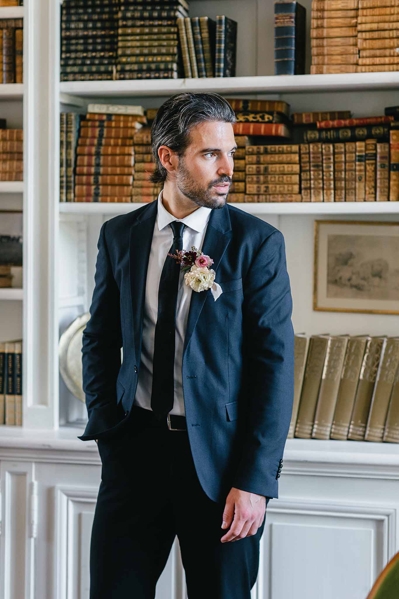 A portrait of the groom standing indoors in front of a white built-in bookcase filled with antique leather-bound books, suggesting a château library or study setting. He is wearing a navy suit with a white dress shirt and a slim black tie, with one hand in his pocket and his gaze directed off-camera to the left. His boutonnière features a cream flower, a small pink bloom, and dark burgundy berry accents. The composition is a three-quarter-length portrait with a classic, formal styling aesthetic.