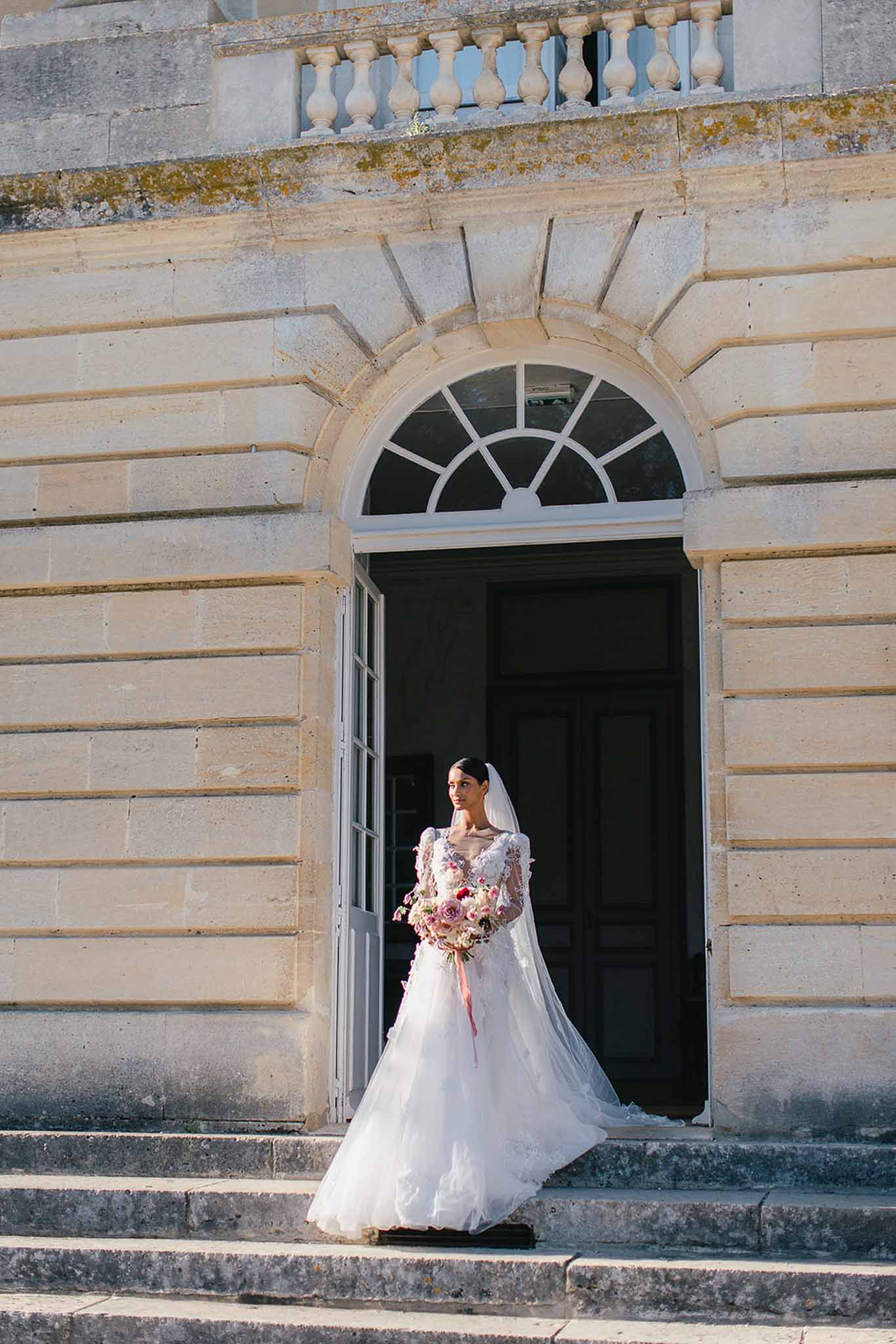 A bridal portrait taken outdoors at a French château, with the bride standing at the base of stone steps in front of a large arched doorway with fanlight window and balustraded upper balcony. The bride wears a long-sleeved white gown with 3D floral appliqué detailing on the bodice and a full tulle skirt, paired with a long cathedral-length veil. She holds a loosely arranged bouquet featuring blush and mauve roses, ranunculus, and trailing pink ribbon. The composition is a full-length portrait framed symmetrically by the classical stone architecture of the building. Potential venue feature image.