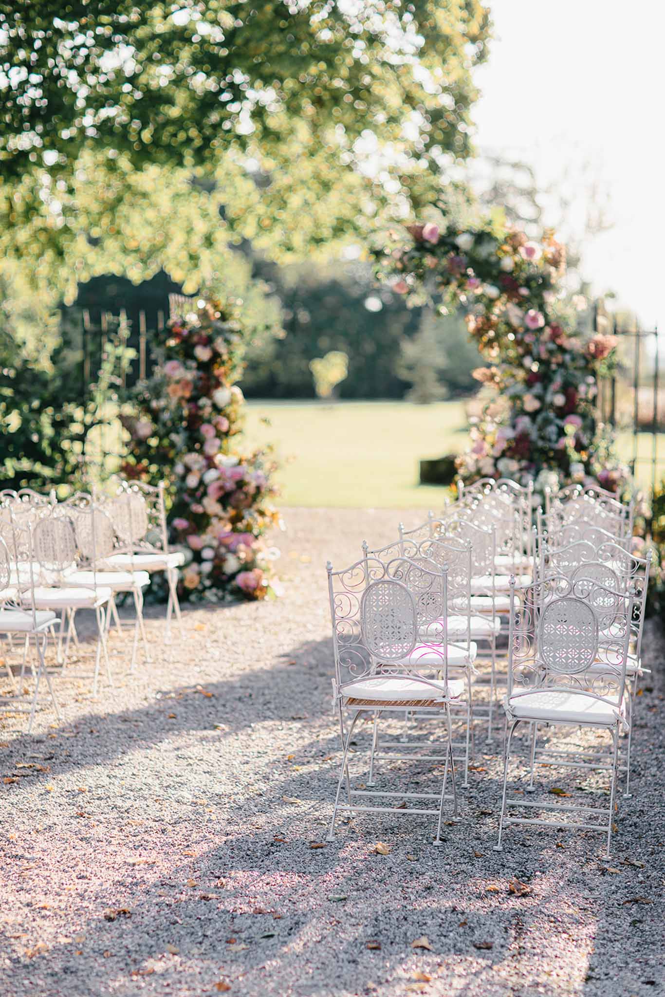 An outdoor wedding ceremony setup on a gravel path, with no people present. Rows of ornate white wrought-iron chairs with white cushioned seats are arranged on either side of a central aisle. At the end of the aisle stands a large circular floral arch decorated with blush pink, dusty rose, burgundy, and cream blooms interspersed with greenery, framing a view of an open lawn beyond. The floral installations extend down to ground level on both sides of the arch, creating full column arrangements. The overall decor palette is romantic and classic, with the white ironwork chairs complementing the soft pink and deep berry tones of the florals. Wide shot taken from ground level looking down the aisle toward the arch. Potential venue feature image.