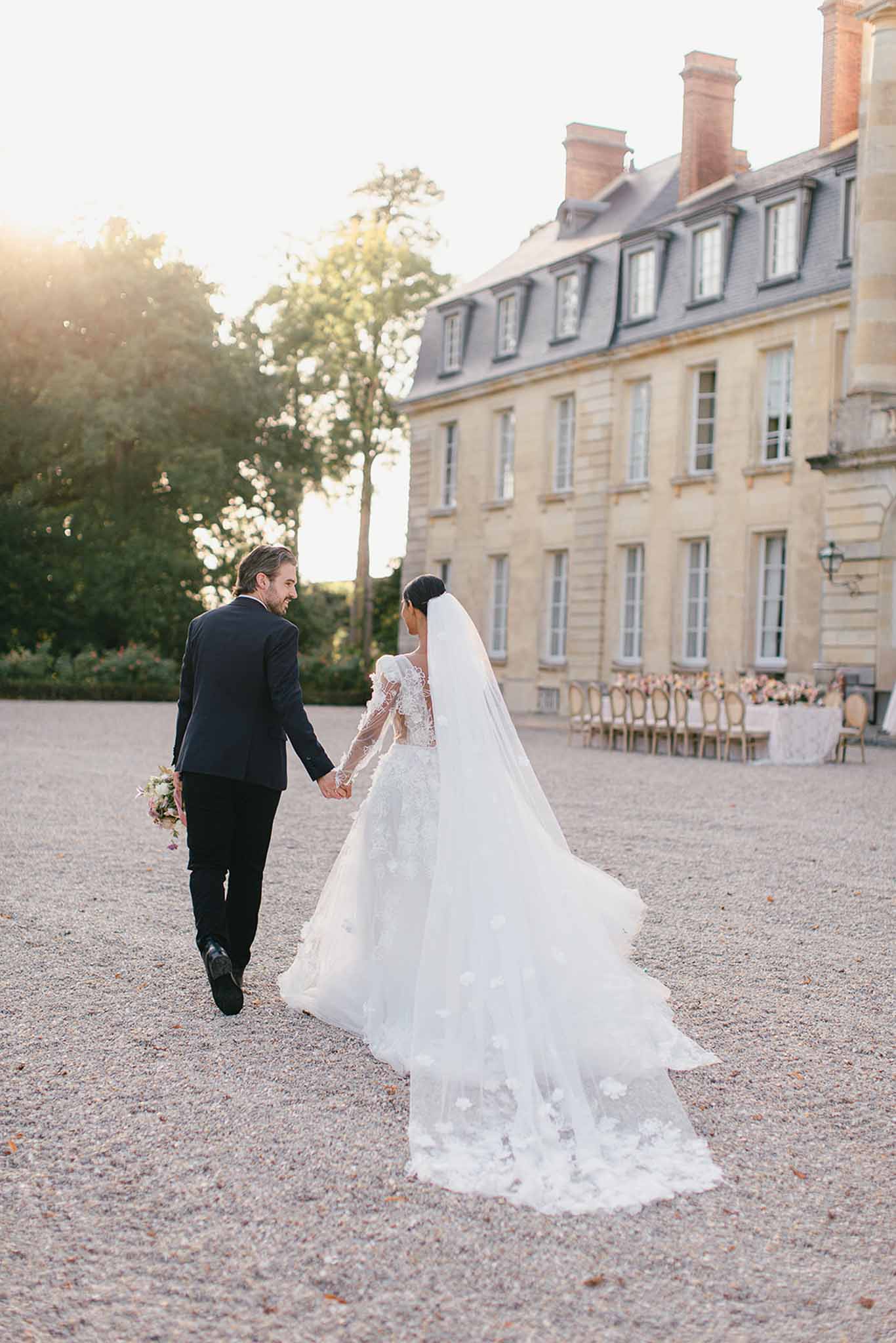 A couple walks hand-in-hand across a gravel forecourt toward a large French château, photographed from behind in a wide portrait shot taken during golden hour. The groom wears a dark navy suit and holds a small floral bouquet with blush and white tones, while the bride wears a white long-sleeve lace gown with three-dimensional floral appliqué detailing and a cathedral-length veil, her hair styled in a low bun. In the background to the right, a long outdoor reception table is set with gold Chiavari chairs and pale pink floral centerpieces arranged in front of the château's classical French limestone façade with mansard roof and brick chimneys. The styling is classic and formal, with warm golden-hour backlight creating a soft glow across the scene. Potential venue feature image.