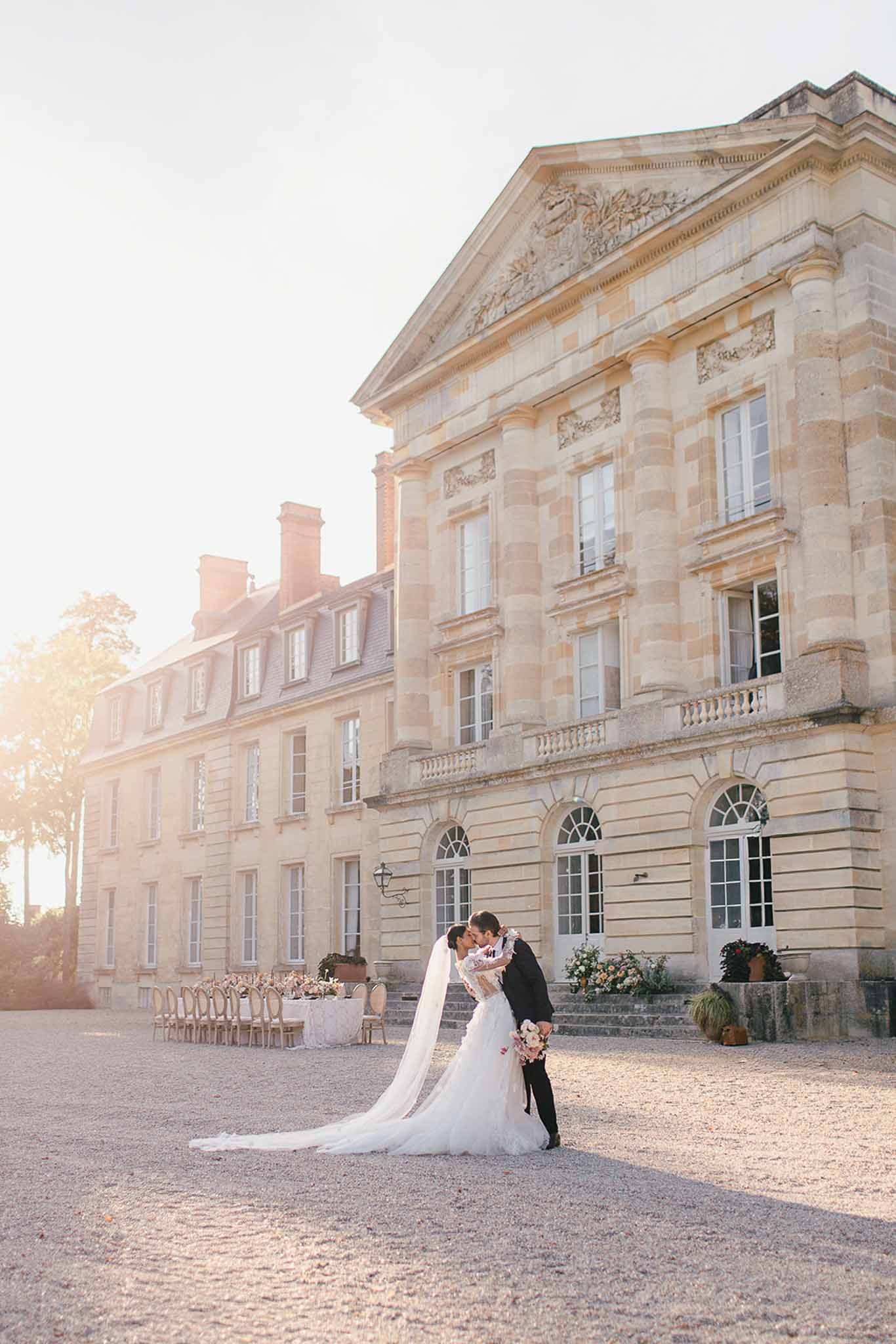 A bride and groom share a kiss on the gravel forecourt of a grand French château during golden hour, with warm low sunlight flooding in from the left side of the frame. The bride wears a fitted ivory lace gown with long sleeves and a long cathedral-length veil that trails across the gravel, and she holds a bouquet featuring blush and ivory blooms. The groom is dressed in a classic black suit. In the background to the left, an outdoor reception table is set with a white linen tablecloth and cross-back chairs, decorated with floral centerpieces in blush and ivory tones, while floral arrangements in similar colors are placed at the base of the château's entrance steps. The shot is a wide full-length portrait that prominently features the neoclassical limestone château facade with its arched ground-floor windows, balustraded balcony, and carved pediment above. Potential venue feature image.