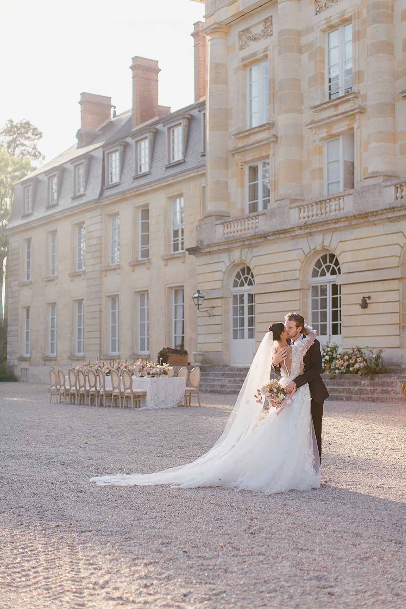A couple shares a kiss during outdoor wedding portraits on the gravel courtyard of a French château, with the full stone facade of the multi-story classical building visible behind them. The bride wears a long-sleeved lace gown with a full tulle skirt and an extended cathedral-length veil, and carries a loose bouquet in muted blush and peach tones; the groom is dressed in a dark navy suit. In the background, a long rectangular reception table dressed in white linen is set along the château facade, lined with gold Louis XVI-style chairs and adorned with floral centerpieces in peach, blush, and warm tones, with additional floral arrangements flanking the entrance steps. The overall styling is classic and romantic, with a warm golden-hour light washing over the scene. This is a wide portrait shot that captures both the couple in the foreground and the reception setup and architecture in full. Potential venue feature image.