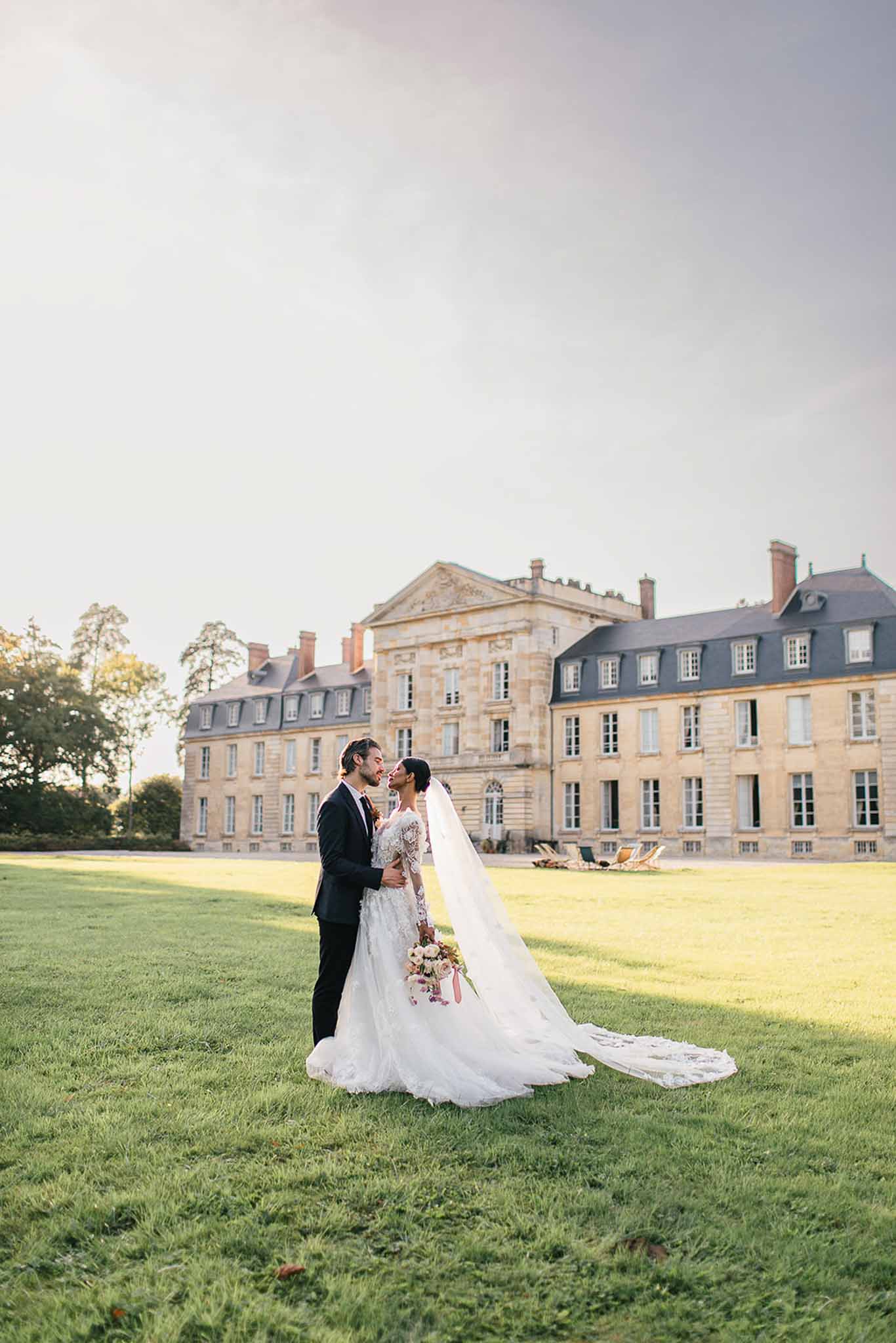 A couple portrait taken outdoors on the grounds of a large French château, shot in warm late-afternoon light. The bride wears a long-sleeved, heavily embroidered ivory gown with a cathedral-length veil and lace-edged train that extends across the lawn; she holds a bouquet of blush, ivory, and burgundy blooms. The groom is dressed in a dark navy suit with a tie, and the two stand close together facing each other as if about to kiss. The wide shot positions the couple in the lower-center of the frame, with the full façade of a classic French château — three stories of dressed limestone with a mansard roof, dormer windows, and a central pediment — filling the background. Potential venue feature image.
