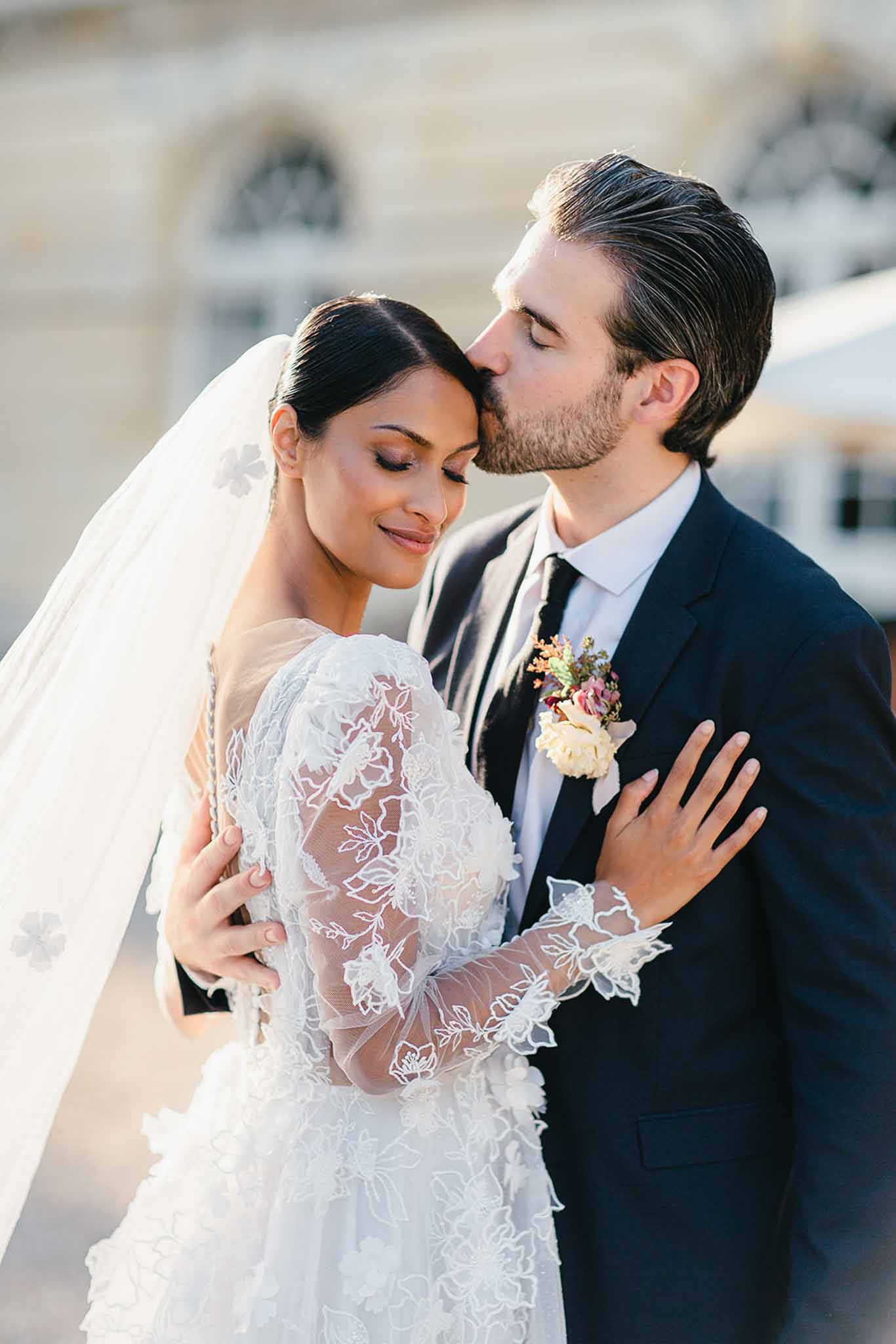 A couple portrait shot outdoors, with the groom kissing the bride on the temple as she smiles with eyes closed. The bride wears a long-sleeve white lace gown with sheer floral-embroidered sleeves and a white tulle veil featuring appliqué floral motifs; her dark hair is pulled back sleekly. The groom wears a navy suit with a white dress shirt, a dark tie, and a boutonnière featuring a cream rose with small burgundy and pink accent blooms. The bride rests her hand on the groom's chest while he holds her waist. The background is softly blurred, showing the pale stone facade of a classical building. The image is a close-up portrait with warm natural light.