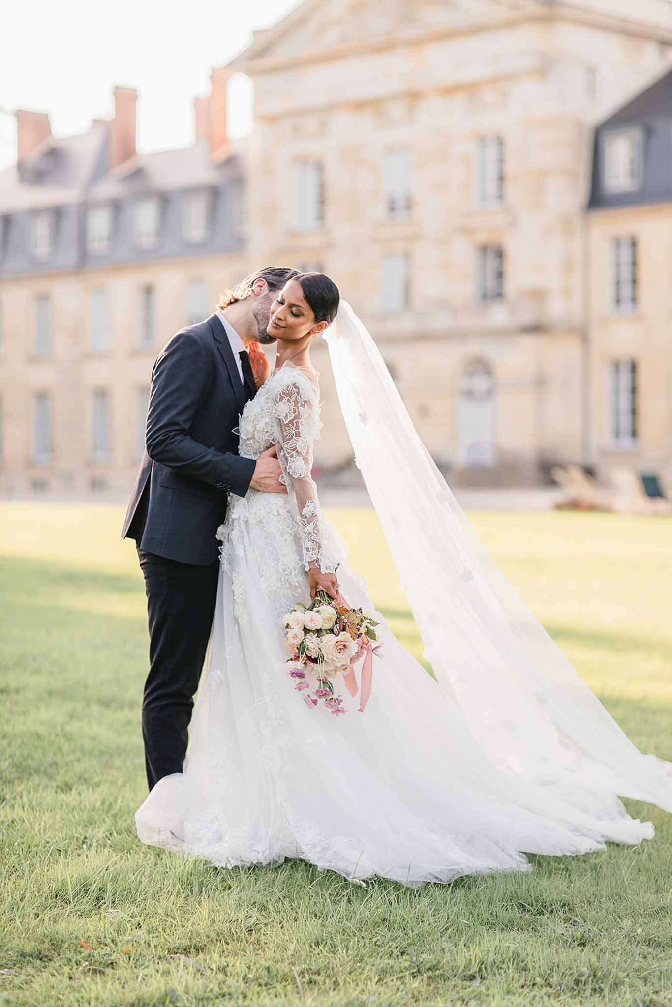 An outdoor couple portrait taken on the grounds of a large French château, with the stone classical-style building visible and softly out of focus in the background. The groom, wearing a dark navy suit with a dark tie, stands behind the bride and kisses her on the temple as she leans into him with eyes closed. The bride wears a full-length white gown with long sheer lace sleeves, three-dimensional floral lace appliqué detailing, a cathedral-length veil that trails across the frame, and holds a loosely gathered bouquet of blush and ivory garden roses with trailing dusty pink satin ribbons. The image is shot as a full-length portrait in warm golden-hour light, with a classic and romantic styling aesthetic.