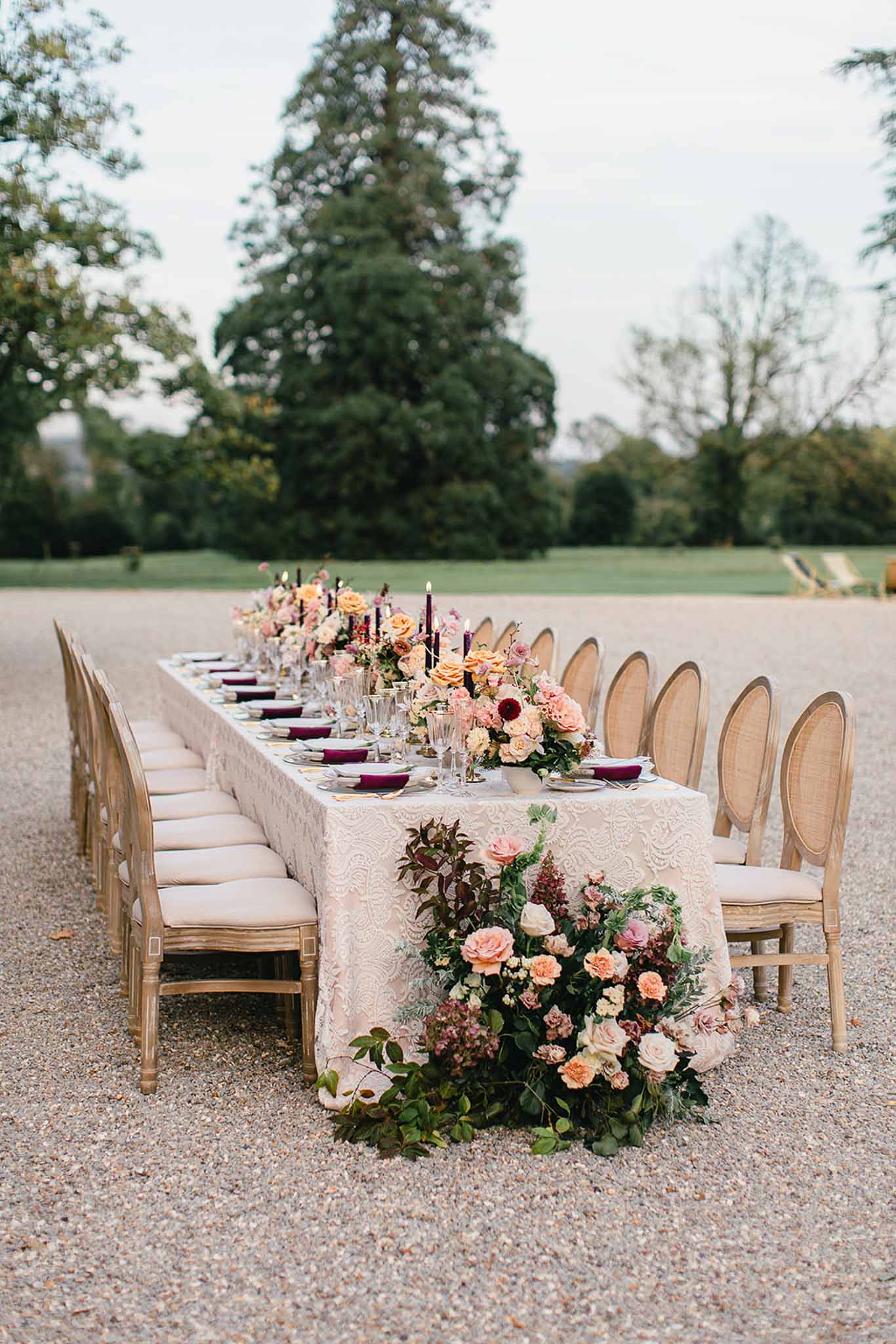 An outdoor wedding reception table styled for a seated dinner, set on a gravel terrace with a large landscaped lawn and mature trees in the background. The long rectangular table is dressed in an ivory embroidered lace tablecloth and lined with Louis XVI-style cane-back chairs in a natural wood finish with cream upholstered seat pads. Place settings feature deep burgundy folded napkins on decorative plates, crystal glassware, and gold cutlery. The floral design runs the full length of the table with arrangements of peach garden roses, blush roses, mauve dahlias, cream blooms, and dark foliage, anchored at the near end by a large ground-level floral installation spilling onto the gravel. Tall dark burgundy taper candles are spaced along the centerpiece. The overall decor palette combines ivory, peach, blush, and deep burgundy in a classic French formal style. Wide shot taken from a low angle along the length of the table.