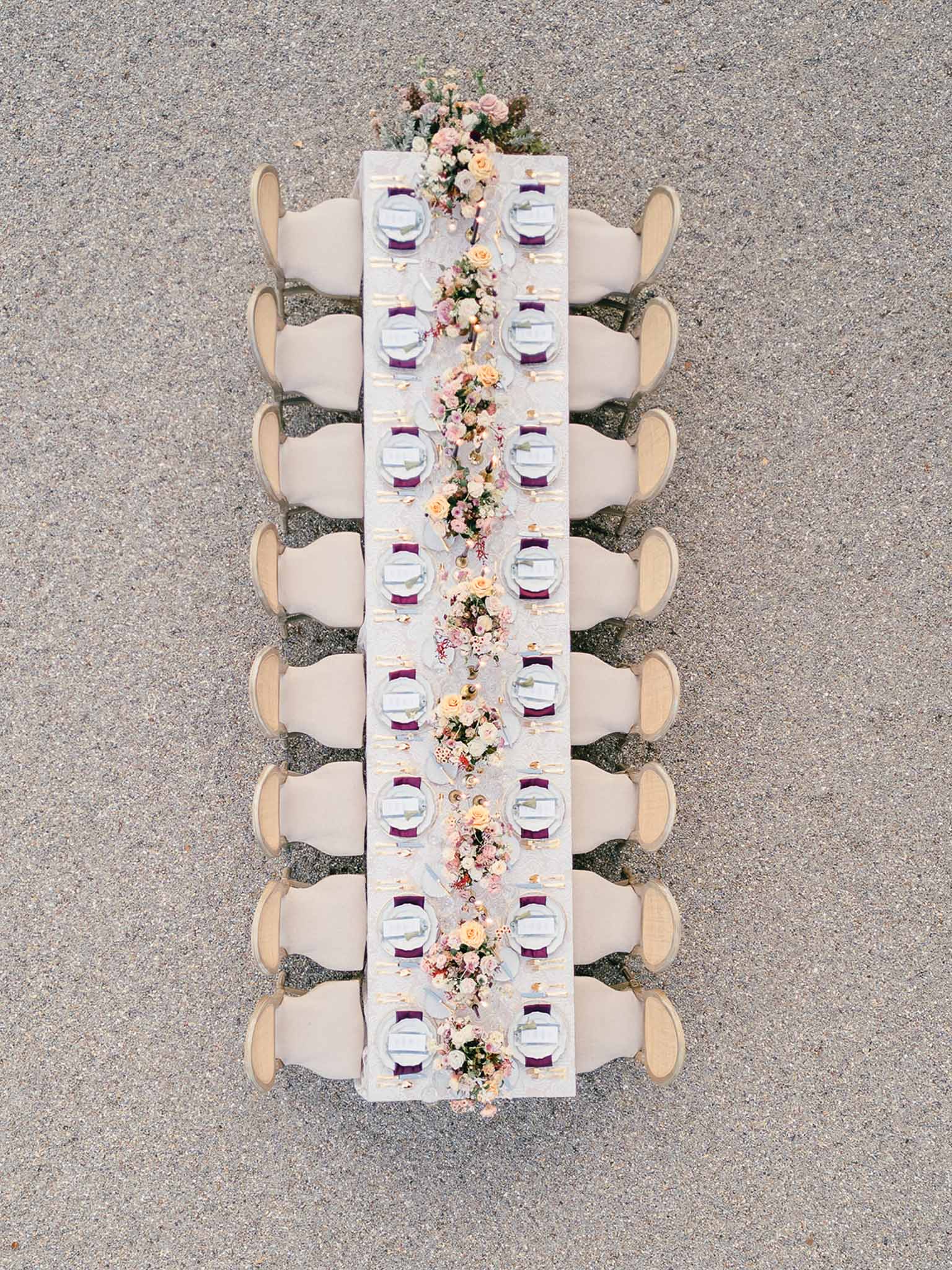 Aerial shot of a long rectangular reception dining table set outdoors on a gravel surface, with no guests present. The table seats approximately 18 people, with cream upholstered chairs arranged along both sides and the ends. Each place setting features a white charger plate with a white folded napkin and a deep plum/eggplant napkin ring or band, accompanied by gold flatware. A continuous floral runner runs the full length of the table, composed of blush pink and peach roses, mauve and dusty rose blooms, small filler flowers in pink and coral tones, and soft greenery. The table linen appears to be white or off-white. The overall decor palette combines cream, gold, plum, and blush tones, reflecting a classic romantic styling approach.