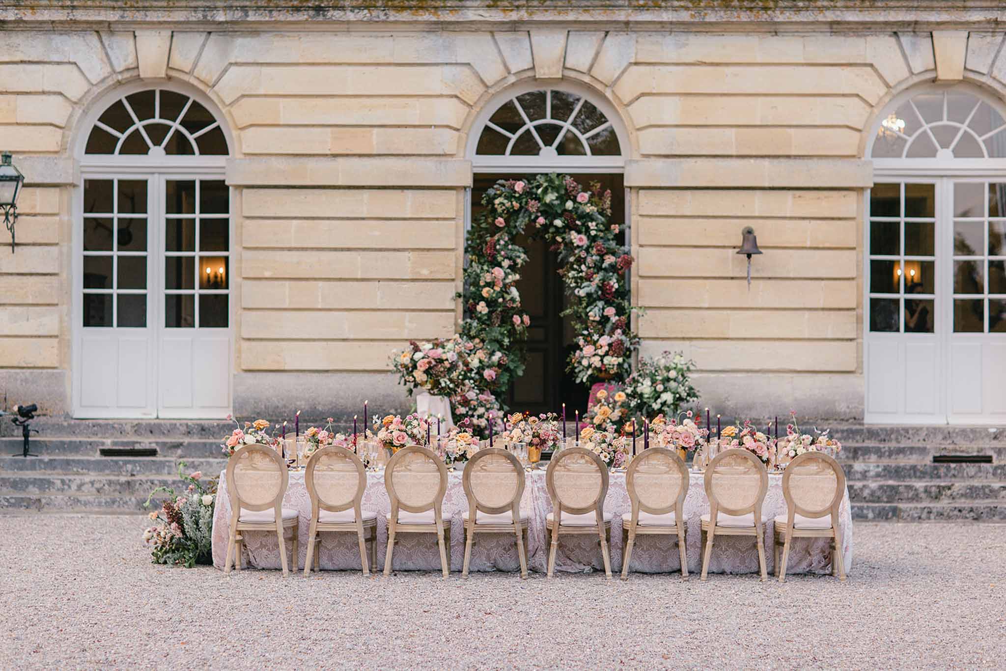 An outdoor wedding reception tablescape set on a gravel courtyard directly in front of a classical French château façade with arched windows and pale limestone architecture. A long rectangular dining table is dressed in a blush and ivory lace or jacquard tablecloth and lined with approximately ten natural wood Louis XVI-style cane-back chairs with blush cushions. The table is decorated with low arrangements of peach, blush pink, coral, and amber garden roses mixed with greenery, interspersed with tall, slender deep plum taper candles and gold bud vases. Centered in the background, a large circular floral arch framing the château's main entrance is constructed from lush greenery, blush and dusty pink roses, burgundy blooms, and trailing foliage, with additional ground-level floral arrangements flanking the doorway. The overall decor palette combines warm blush, peach, amber, and deep plum tones in a classic French formal style. Wide symmetrical shot taken straight-on from ground level. Potential venue feature image.