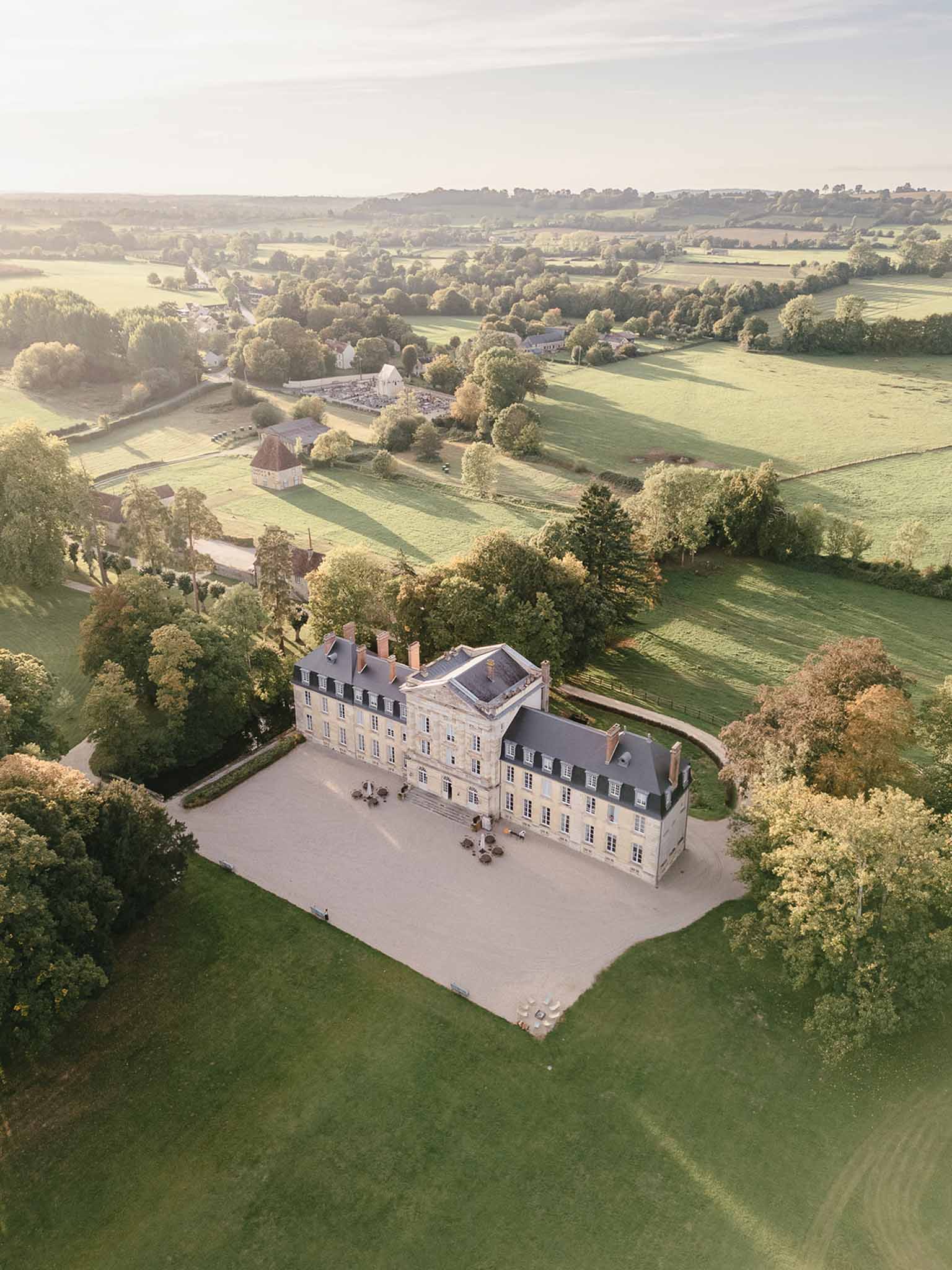 Aerial drone shot of a large French château with cream-colored stone façade, grey slate roof, multiple chimneys, and symmetrical multi-pane windows across three stories. The building features a central projecting section and is flanked by curved stone walls leading to outbuildings. A gravel forecourt in front of the château holds several outdoor seating arrangements — what appear to be round tables with white umbrellas or parasols. The property includes a formal driveway approach, extensive lawns, and is surrounded by mature trees. Further outbuildings, including a structure with a terracotta-tiled turret roof, are visible in the mid-ground, along with what appears to be a set-up event space with white furniture arrangements to the upper left. The wider landscape shows rolling countryside with fields and hedgerows stretching to the horizon. Potential venue feature image.