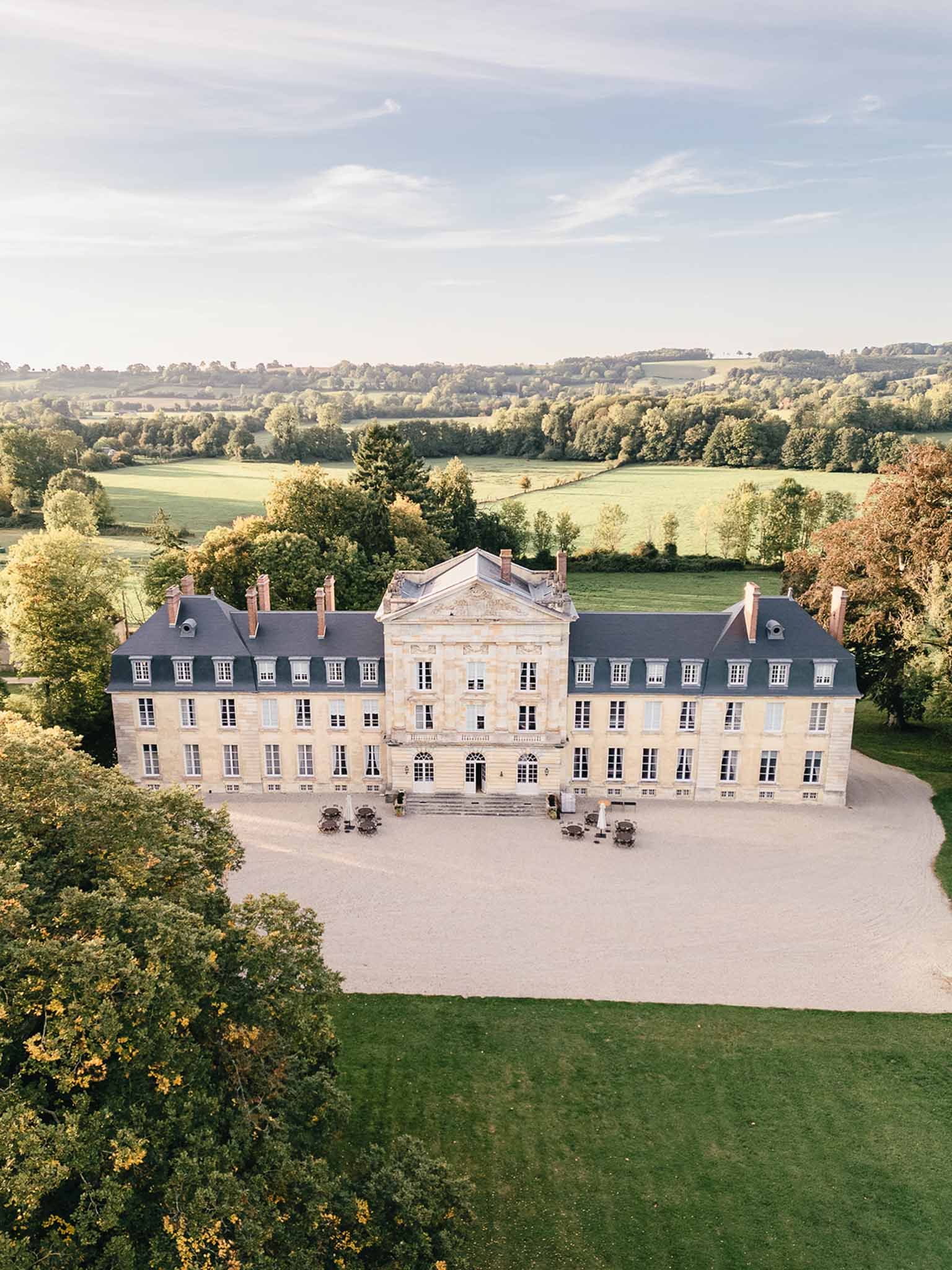 Aerial drone shot of a large French classical château featuring pale limestone façade, dark slate mansard roofs with multiple chimneys, and a central pedimented pavilion with ornate carved stonework above the main entrance. The building spans three stories with symmetrical rows of white-shuttered windows across its full width. A wide gravel forecourt sits directly in front of the entrance, with several outdoor seating arrangements — round tables with chairs — positioned on either side of the main steps. The château is surrounded by manicured lawn, mature trees showing early autumn foliage in yellow and green tones, and open countryside with rolling fields and treelines extending into the background. No people are visible in the image. Potential venue feature image.