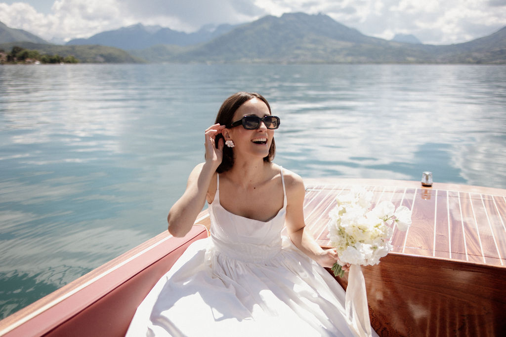 A bride sits aboard a classic wooden motorboat with red leather upholstery on a mountain lake, laughing and adjusting her pearl cluster earring. She wears a white spaghetti-strap wedding dress with a gathered skirt, black rectangular-frame sunglasses, and holds a bouquet of white peonies and sweet peas with a white ribbon wrap. The portrait is shot from the bow of the boat at a slight downward angle, with the lake and mountain range visible in the background. The overall styling is modern and relaxed, mixing a classic bridal look with casual accessories.