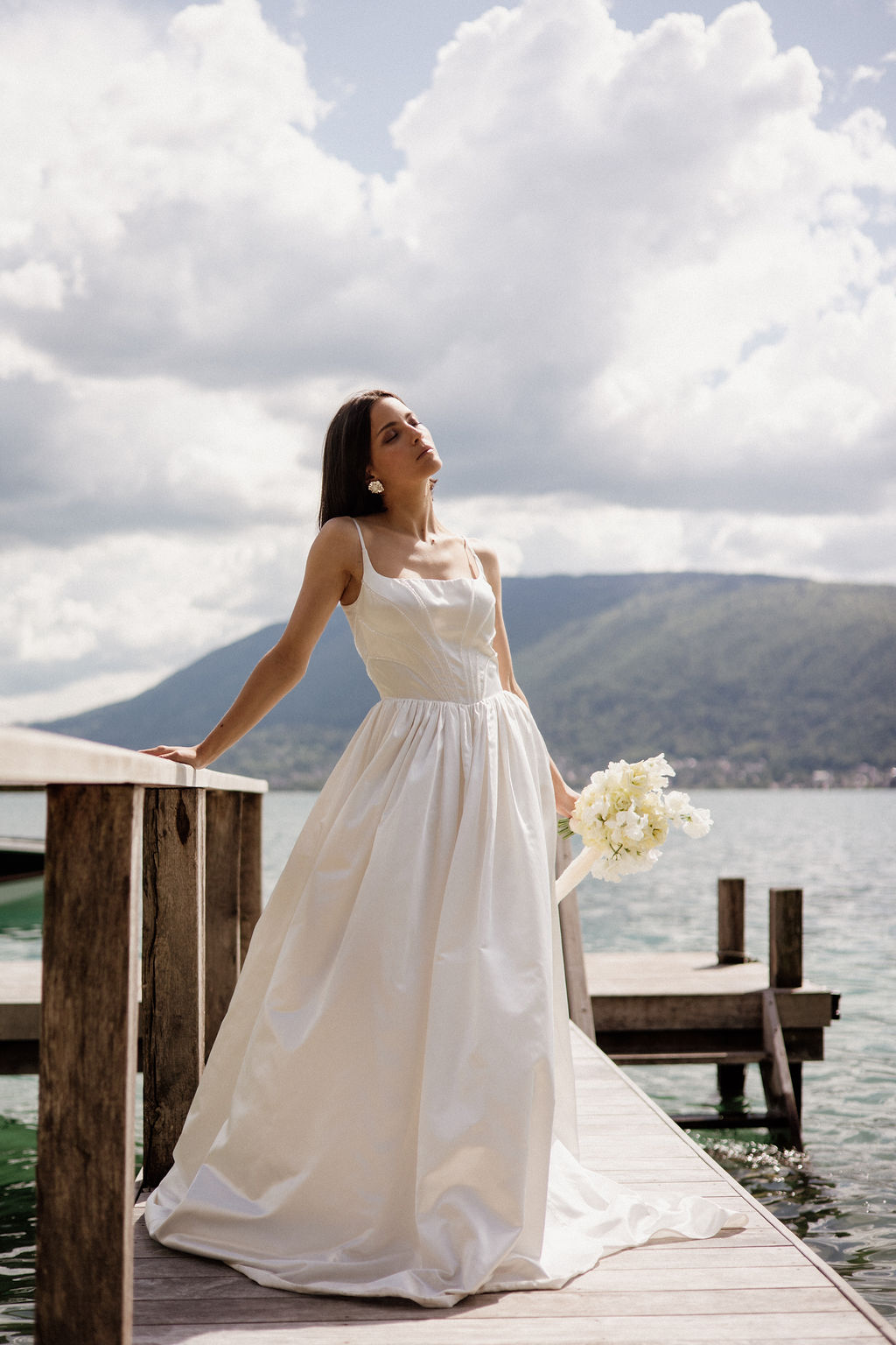A bridal portrait taken outdoors on a wooden lakeside dock, with a mountain lake and rolling hills visible in the background. The bride, a dark-haired woman, stands alone in a full-length white gown with thin spaghetti straps, a fitted bodice, and a full gathered skirt with a short train; she wears statement gold floral drop earrings and holds a compact bouquet of white sweet peas or ranunculus loosely at her side. Her head is tilted upward with eyes closed in a posed, editorial style. The composition is a medium-full portrait shot taken at slight low angle, giving prominence to the dress silhouette against the open water and sky backdrop, in a clean, modern bridal style.