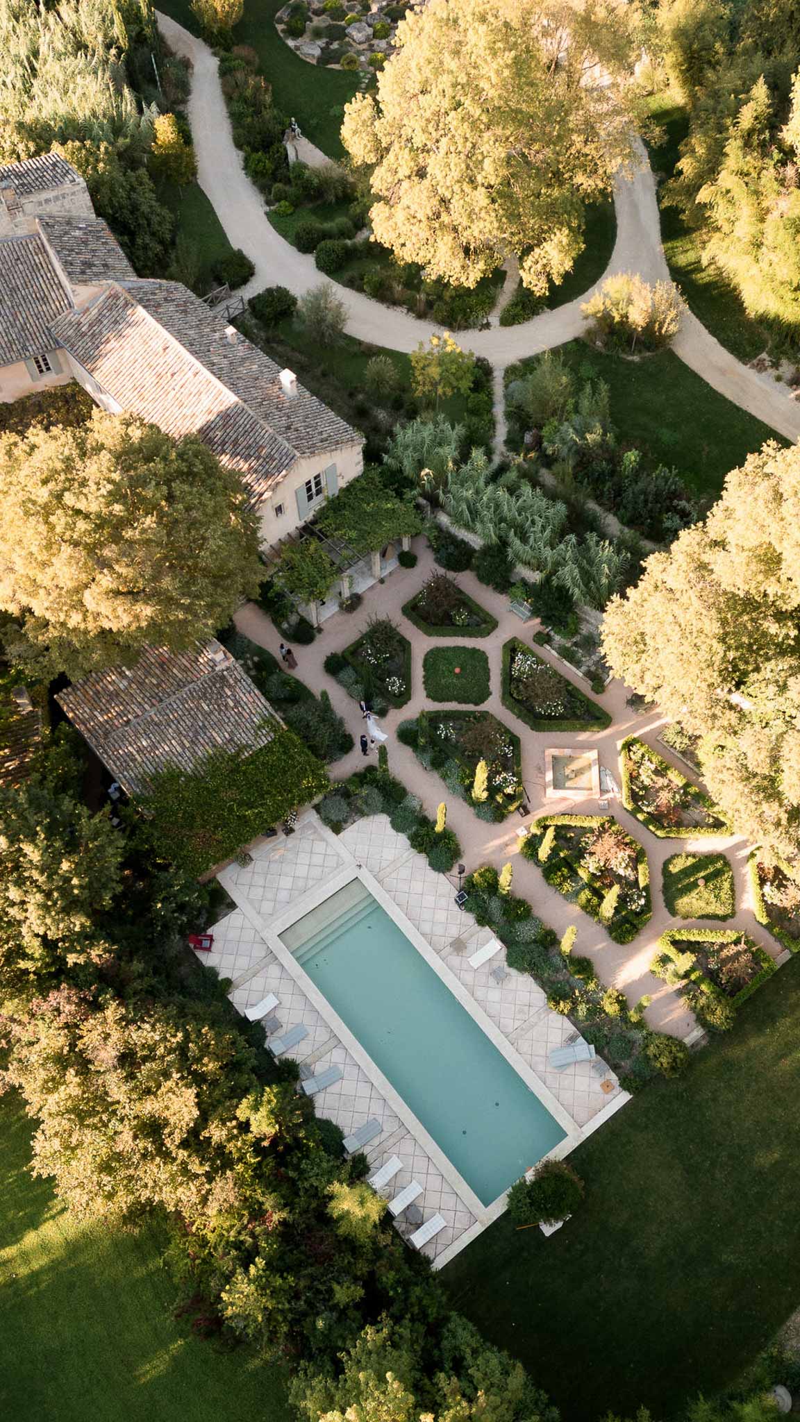 An aerial drone shot looking straight down over a French country estate, showing the property's grounds in full. The venue features a large rectangular lap pool with a pale blue-green tiled surround and white sun loungers arranged along its edges, positioned at the lower portion of the frame. Above the pool, a formal parterre garden is visible with geometric boxwood-edged beds, gravel pathways, and planted borders containing white flowering plants. A pergola structure connects to the main building, which has terracotta-tiled roofing typical of Provençal architecture with stone-colored rendered walls. Curving gravel driveways wind through the upper portion of the grounds. Two or three small figures in formal attire, including what appears to be a figure in white, are visible walking through the garden area near the parterre, suggesting a wedding is taking place on the property. Potential venue feature image.