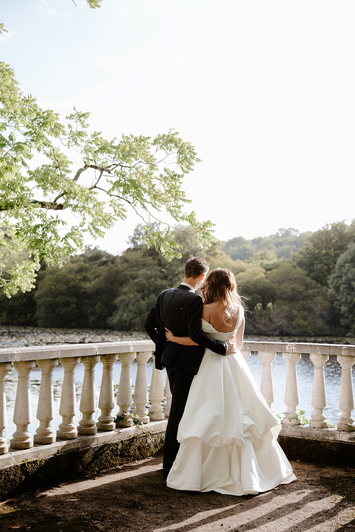 A couple portrait taken outdoors on a stone balustrade terrace overlooking a lake or river, shot from behind. The groom wears a black suit and has his arm around the bride, who wears a white strapless ballgown with a low open back and tiered full skirt. The two are turned away from the camera, facing the water. Small trailing greenery is placed along the base of the white stone balustrade. The image is a full-length wide portrait with strong backlighting creating a bright, hazy quality. The classic French château-style stone balustrade and lakeside setting suggest a formal estate venue.